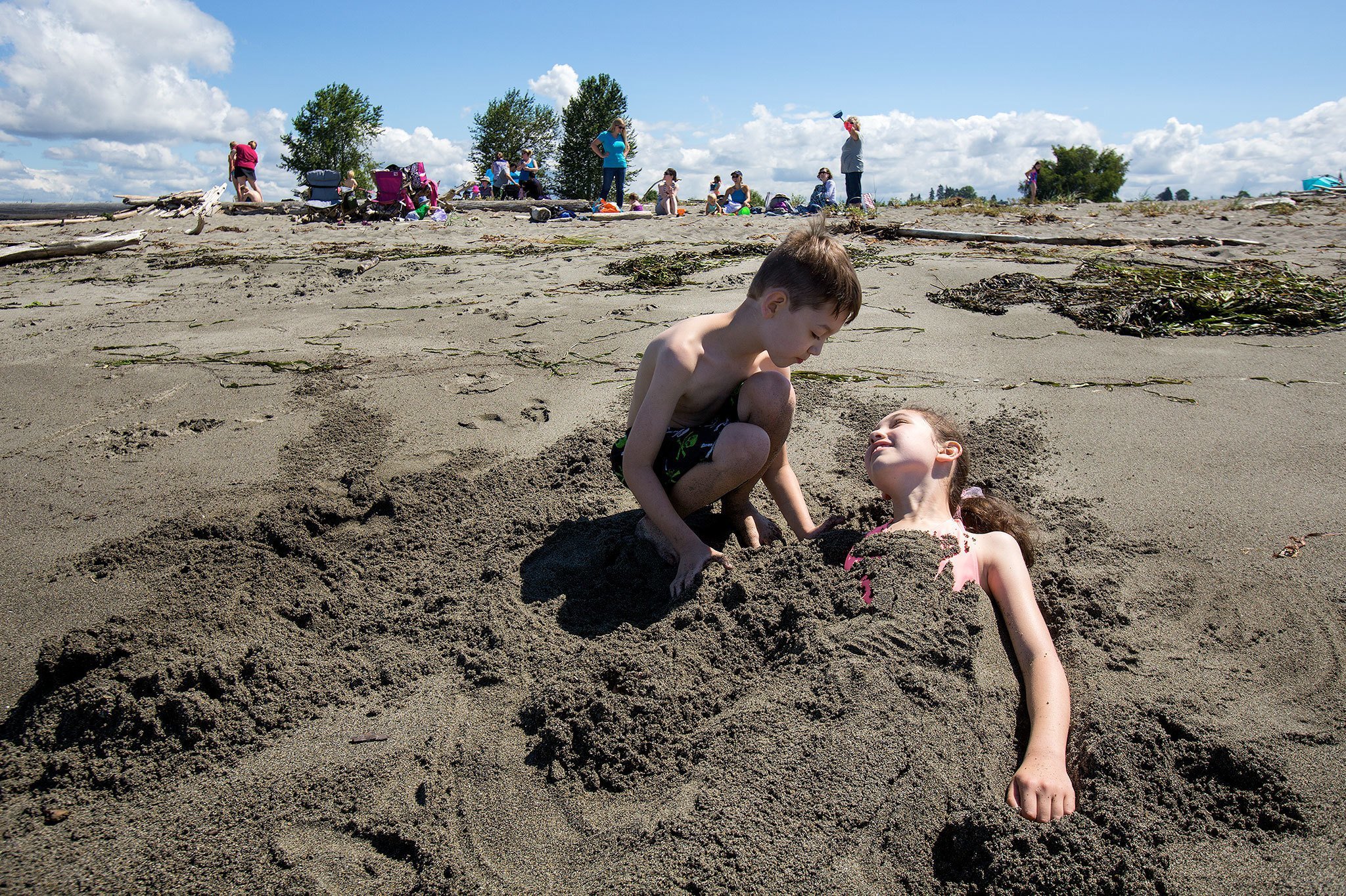 Isabelle Shaw, 10, looks at her brother, Keoki, 8, as he buries her in the sand on Jetty Island after the ferry began operations for the summer on Wednesday in Everett. (Andy Bronson / The Herald)