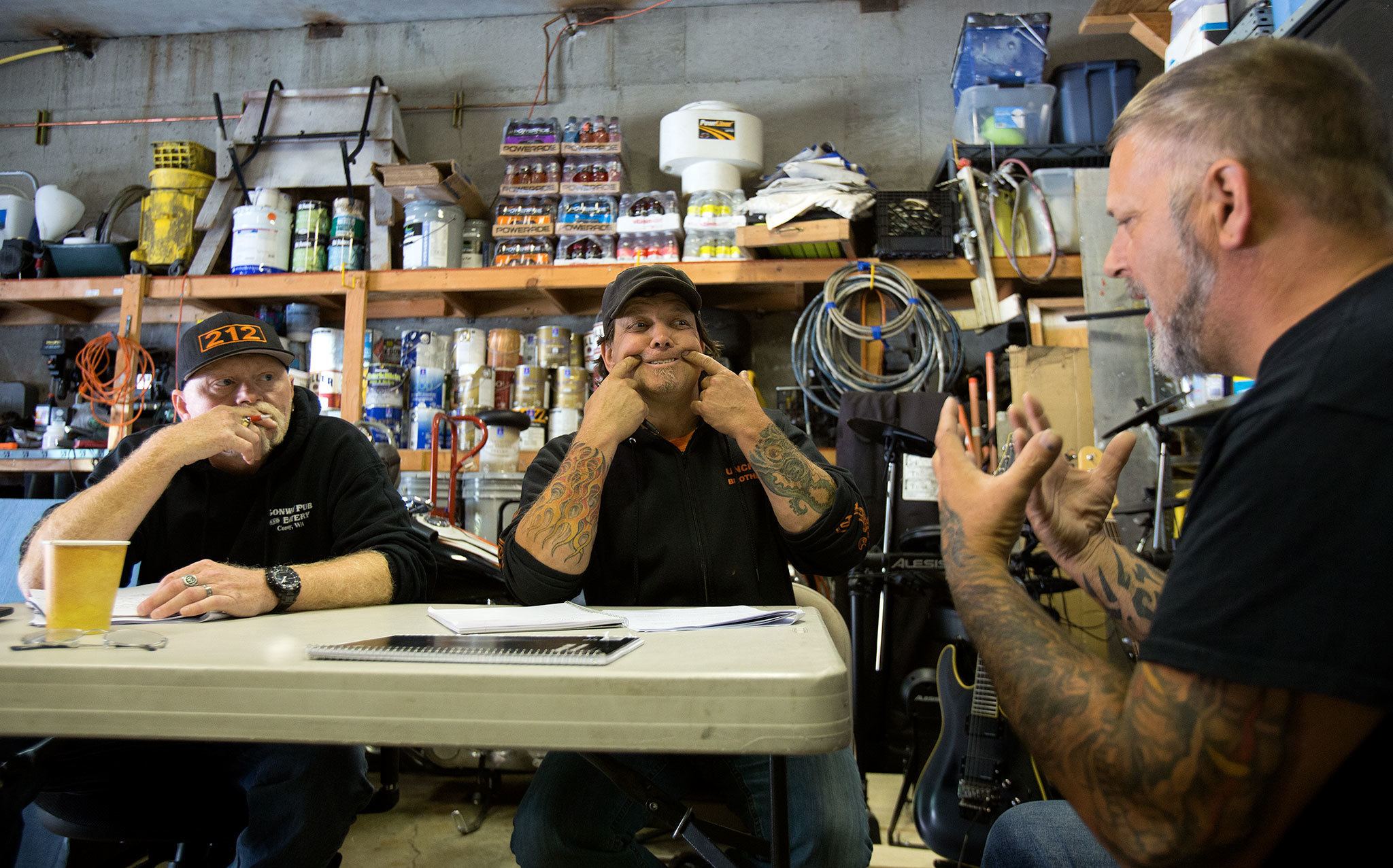 Practicing life coach interviewing skills, Jerry Remington puts a smile on his face, showing how a better spiritual life looks “smiling more” to Chris Hagins as they practice in Remington’s garage on July 10 in Everett. Watching and critiquing is Steve “Redd” Arnold (left). Remington, the president of the Everett chapter of the Unchained Brotherhood, a clean-and-sober motorcycle club, is leading the group toward becoming certified in life and recovery coaching. (Andy Bronson / The Herald)