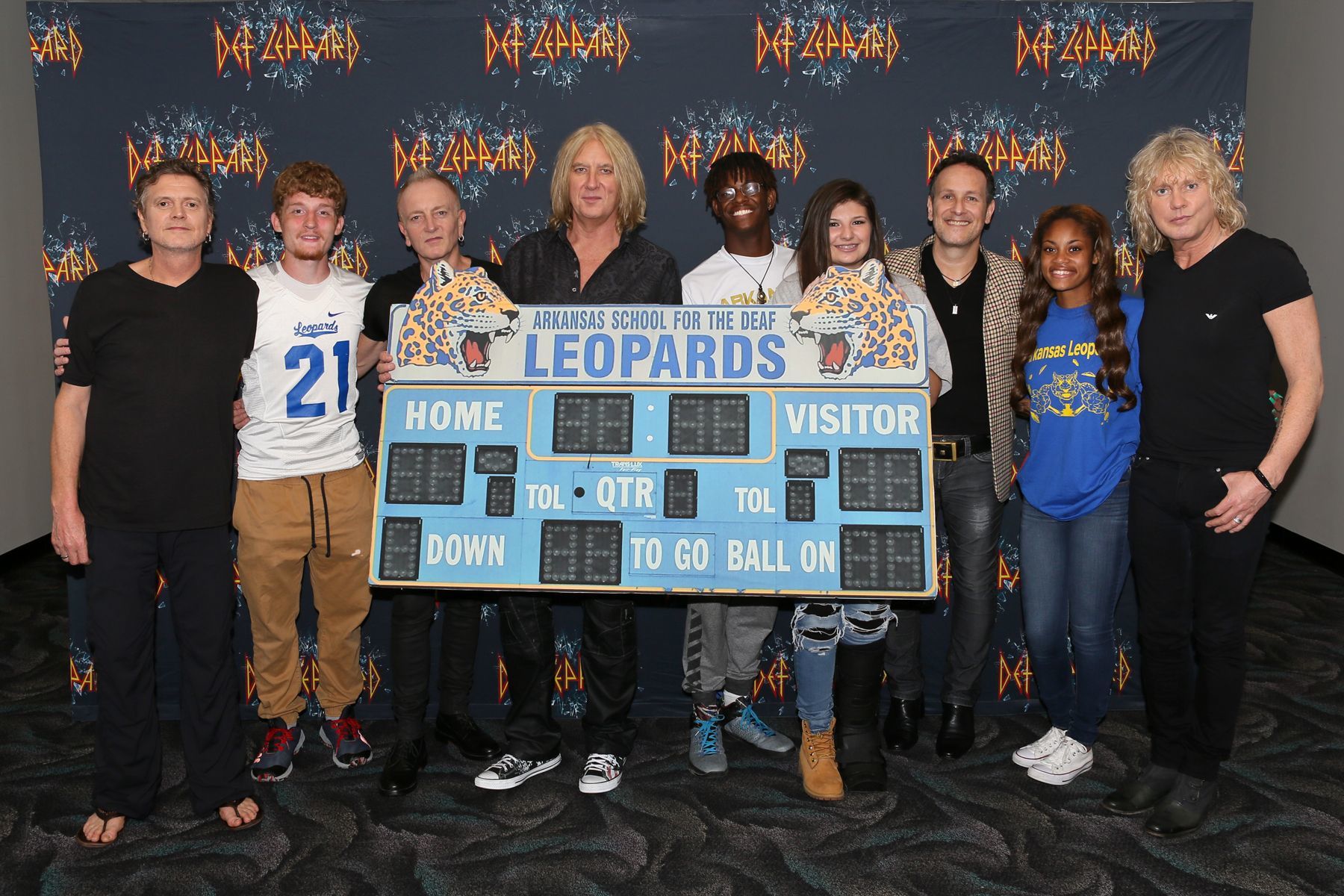 From left, Def Leppard drummer Rick Allen, student Stephen Cathcart, guitarist Phil Collen, lead singer Joe Elliott, student Henry James, student Alex Gossett, guitarist Vivian Campbell, student Jewel Brandon and bassist Rick Savage pose with a replica of the scoreboard from the Arkansas School for the Deaf, before a concert in North Little Rock, Ark.