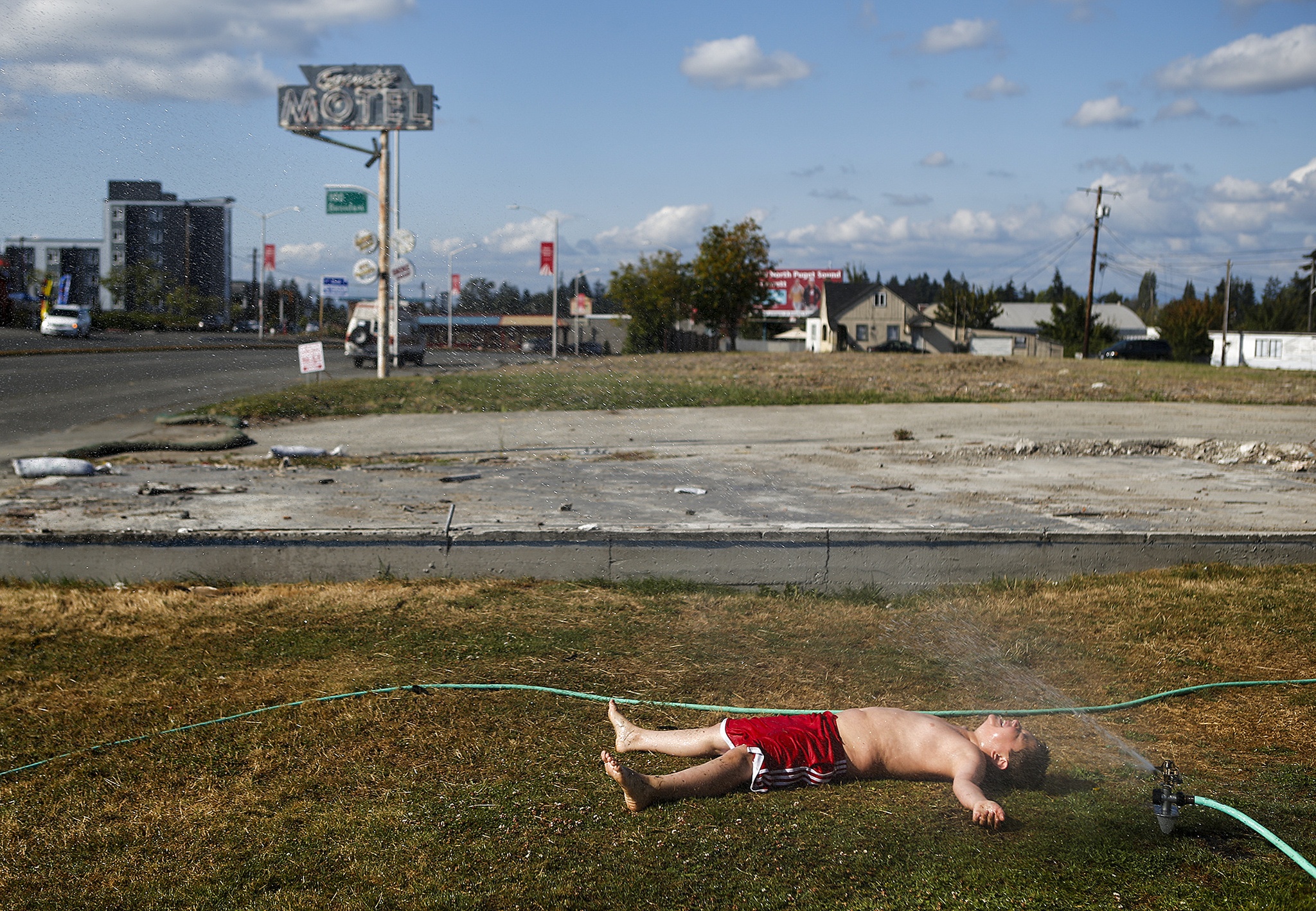 Sylas Westgard, 9, keeps cool with a sprinkler as he lays in the grass near his family’s home on north Broadway in Everett on Aug. 3. The old “Everett Motel” sign is all that remains in the empty lot next to Westgard’s home. (Ian Terry / The Herald)