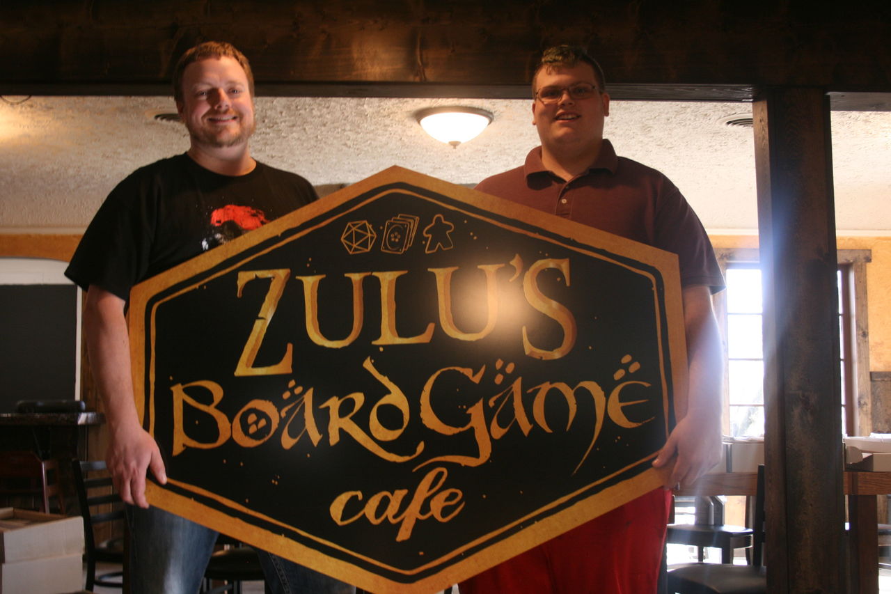 Zulu’s Board Game Cafe owner Matt Zaremba (left) and Kaleb Muse show the sign for the business that’s opening at 10234 Main St. in downtown Bothell.