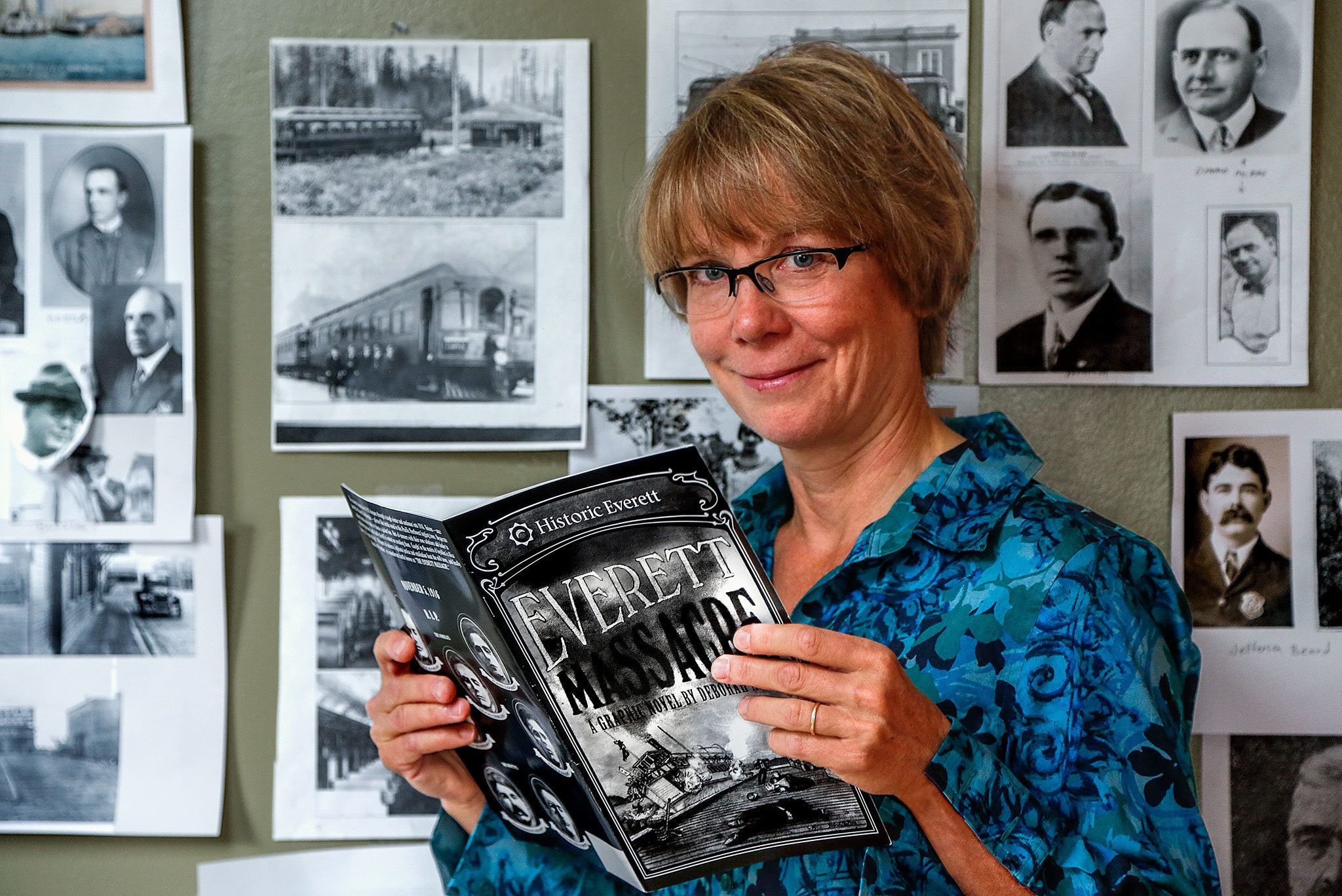 In her home in Everett, artist Deb Fox holds a copy of a new graphic novel she created based on the Everett Massacre. Photocopies of historic photographs hang on her studio wall behind her. She created the art for the graphic novel using charcoal. (Dan Bates / The Herald)