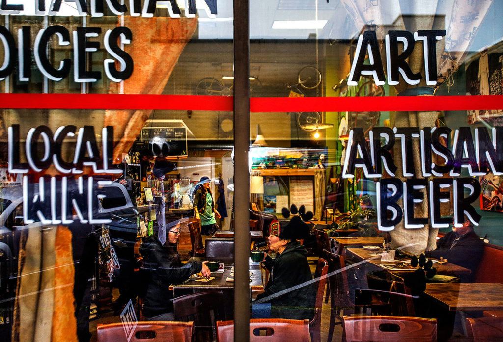 In a cozy atmosphere, partially visible through painted signs and street reflections on the front window panes, Janet Hada (left front table) and her sister, Susan Hada, who work for the U.S. Forest Service in Everett, enjoy coffee Tuesday morning at Cafe Zippy on Wetmore Avenue, where they are regular customers. Rich Horton works on his computer at a table off to the right. Marilyn Rosenberg, owner (in green apron, back), is preparing to move the restaurant/coffee-house from 2811 Wetmore Ave. to the former home of Under the Red Umbrella. That cafe, at 1502 Rucker Ave. in north Everett, closed in 2015. (Dan Bates / The Herald)
