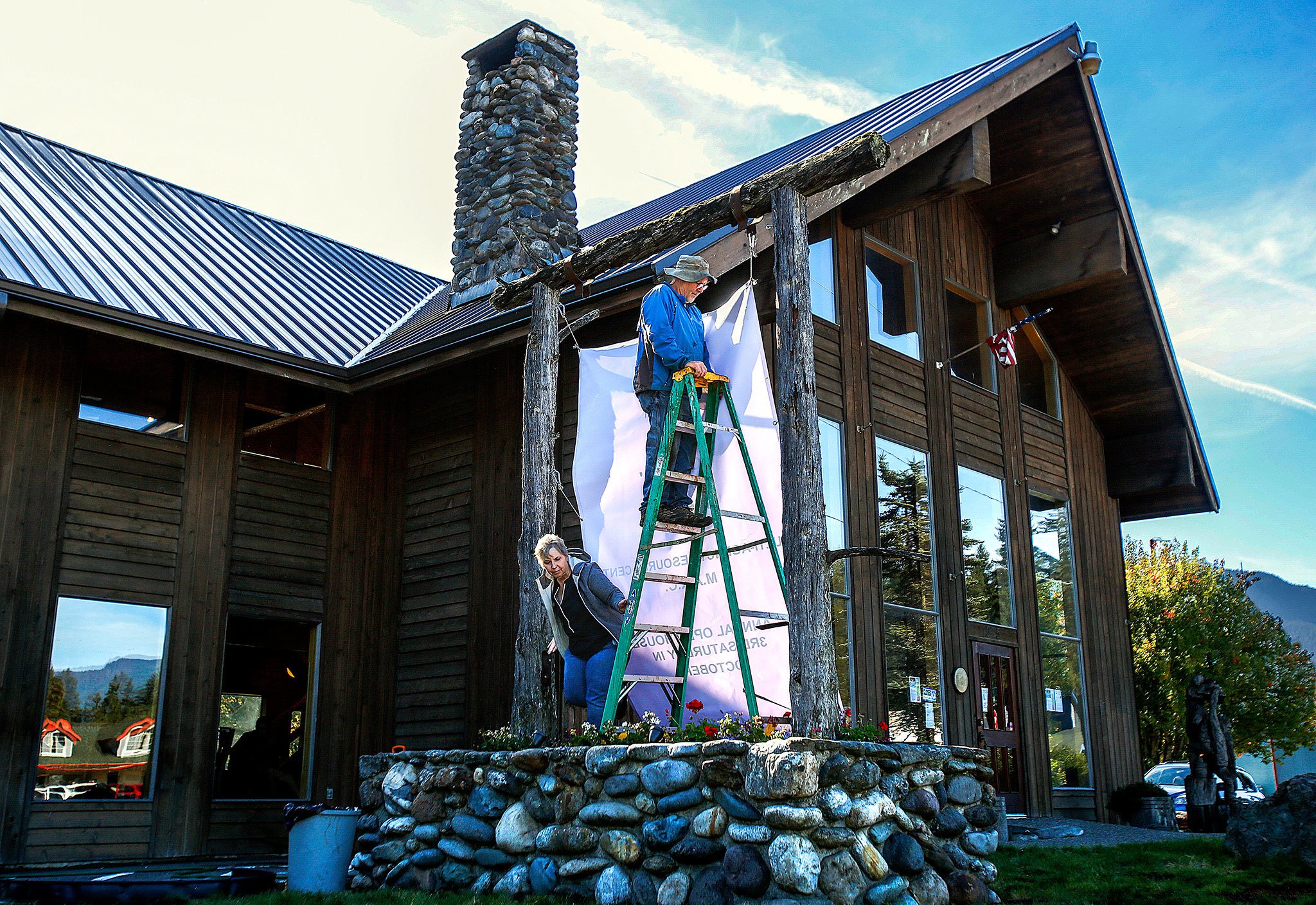 Executive Director and assistant sign hanger, Wyonne Perrault, slips around the bottom of the lader to help her husband Joe install a large banner outside the new North Counties Family Services facility in Darrington Wednesday. (Dan Bates / The Herald)                                Executive Director Wyonne Perrault helps her husband, Joe, install a large banner outside the new North Counties Family Services facility in Darrington in October. (Dan Bates / The Herald)
