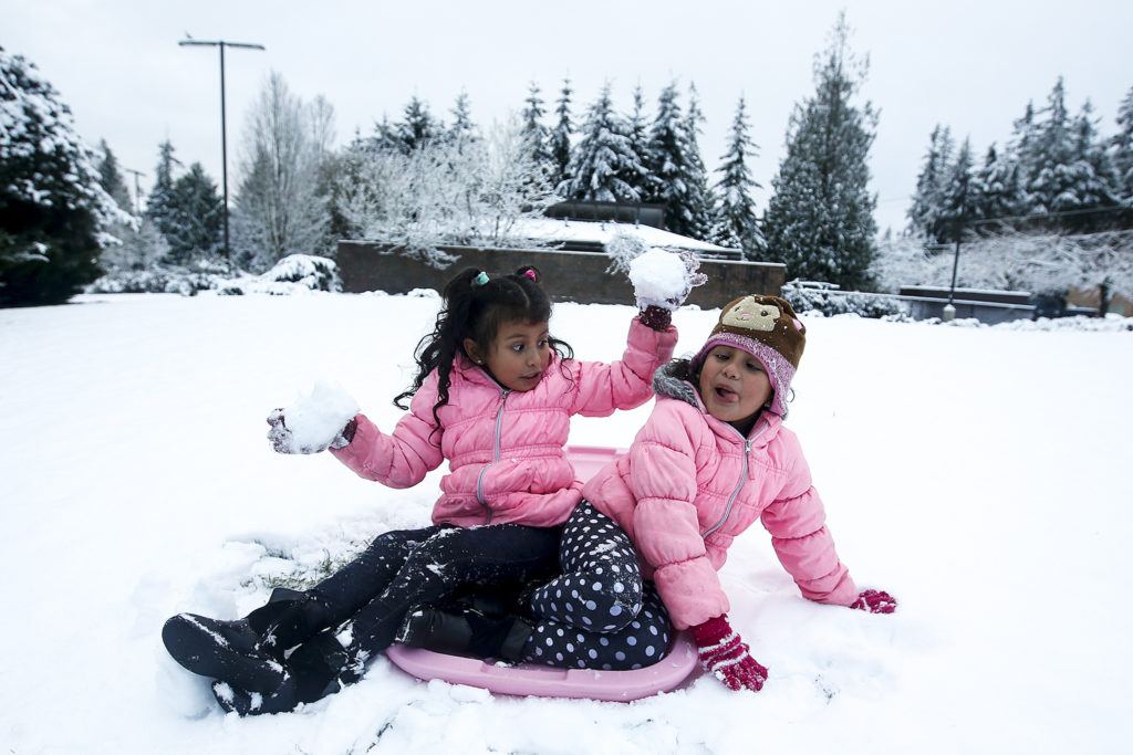 Jennifer Ardon (left) grabs two fistfuls of snow for her twin sister Stephanie while waiting for the bus with their dad Fredi (not pictured) in Lynnwood on Friday morning. (Ian Terry / The Herald)
