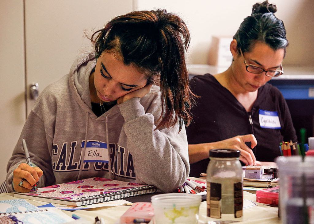Ella Estlund (left) writes with tiny letters in the spaces around graphic designs she created using a fine-point pen. Cynthia Gahan uses rubber stamps to apply the letters of words in her journal. (Dan Bates / The Herald)
