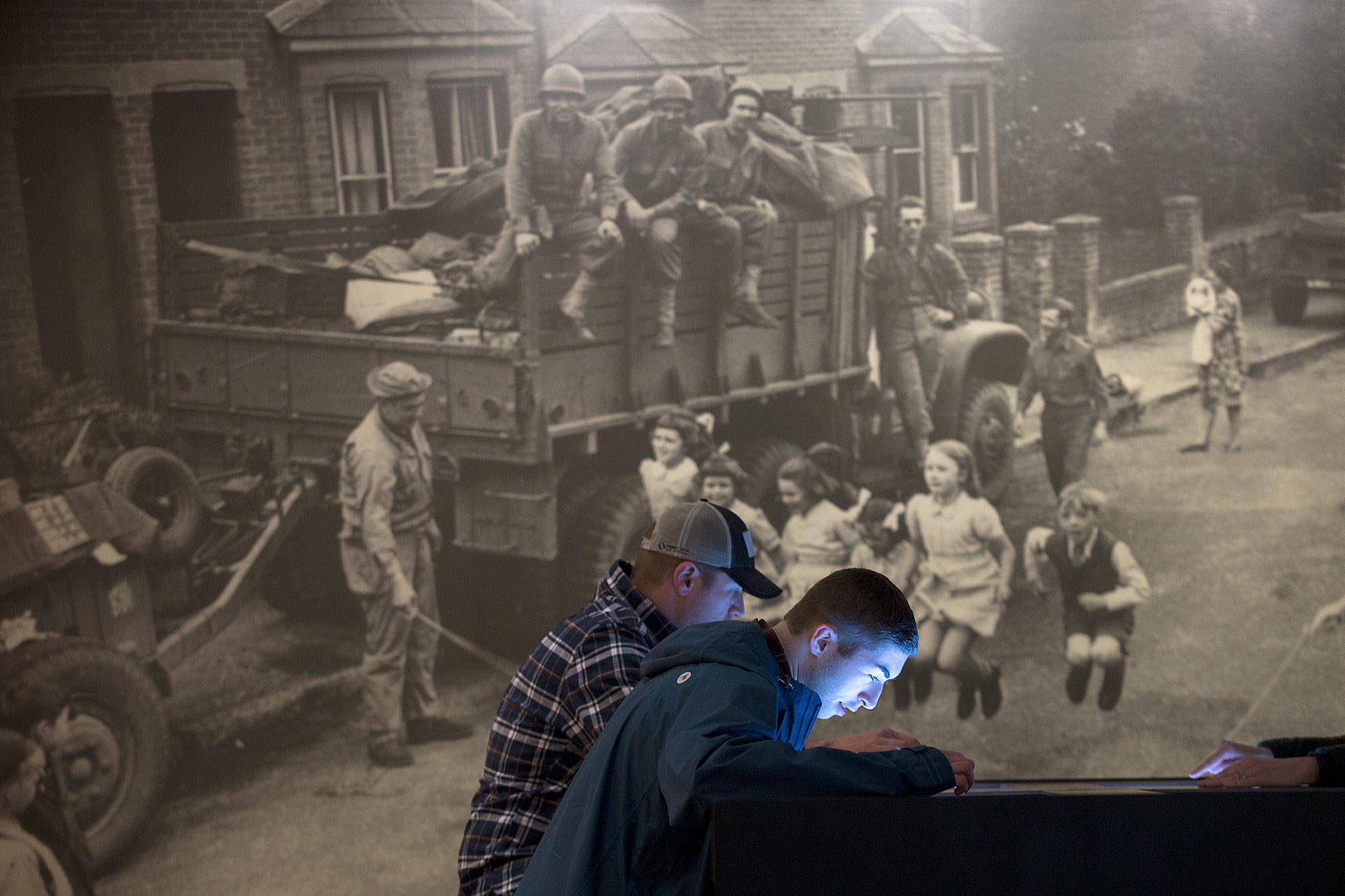 A mural shows soldiers watching children jumping rope as Hunter Thompson, 17, front, listens to war stories next to his father, Jason, in the Flying Heritage & Combat Armor Museum’s new exhibit, “Why War?” on Thursday in Everett. (Andy Bronson / The Herald)