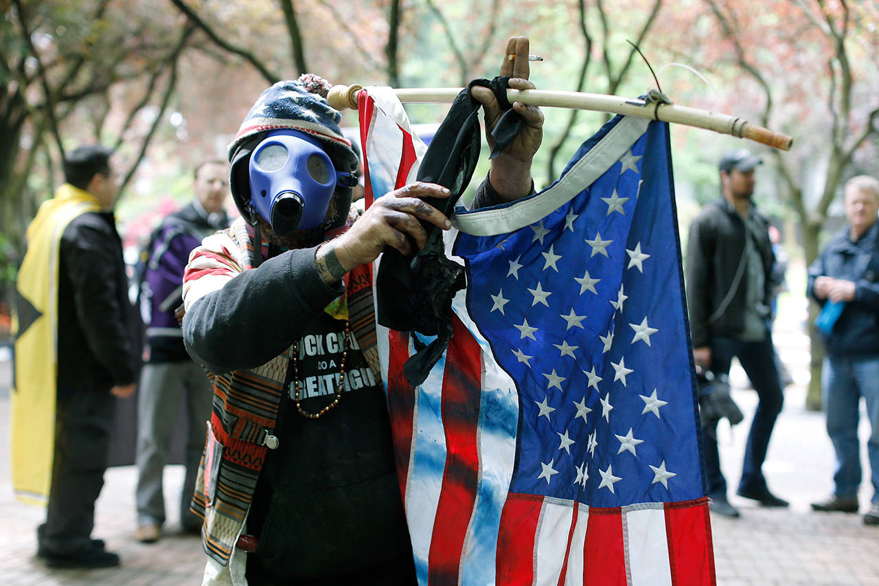 John Durano, who says he flew in from Los Angeles to participate and demonstrate his white pride, wears a gas mask and waves an American flag Sunday during a demonstration near City Hall and adjacent parks in Portland, Oregon. Crowds swelled to several thousand as demonstrators from varying groups converged in downtown Portland. (Beth Nakamura/The Oregonian via AP)