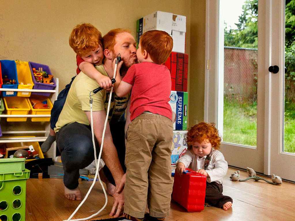 In their basement playroom Thursday morning, Alex Clawson, 32, grants a kiss to son Atley, 3, while son Willem, 4, climbs up his back and 20-month-old daughter Seirsha tries to open a plastic container. (Dan Bates / The Herald)
