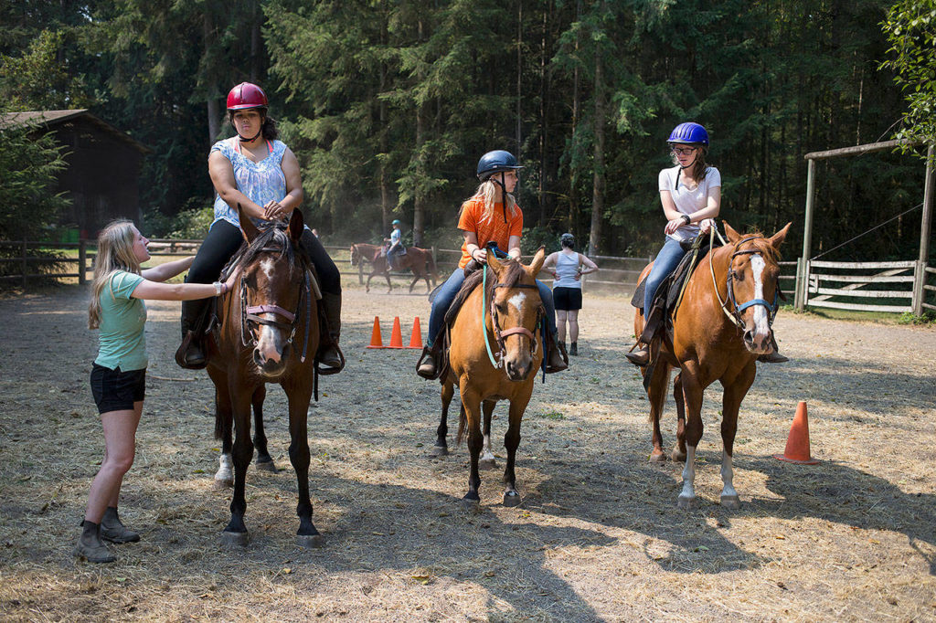 Horse riding staff member Bella Harris (left) talks with Michou Mayer during a riding class at Camp Killoqua on Aug. 2 in Stanwood. McKenzie Levi and Sammi Sutton (right) wait their turn around the arena. (Andy Bronson / The Herald)
