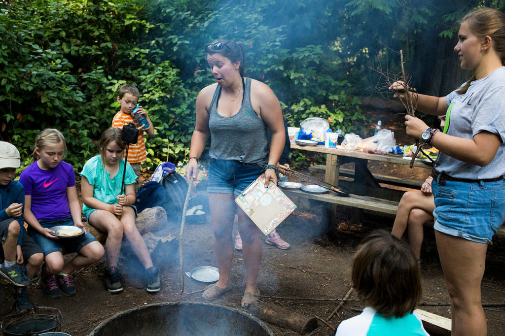 Smoke gets into the eyes of counselor Lauren Jones, aka “Socks,” as she tries to get a fire going so day campers can make pie iron pizzas at Camp Killoqua on Tuesday in Stanwood. Instead of proper names, camp leaders and volunteers are referred to by nicknames throughout the week. (Andy Bronson / The Herald)
