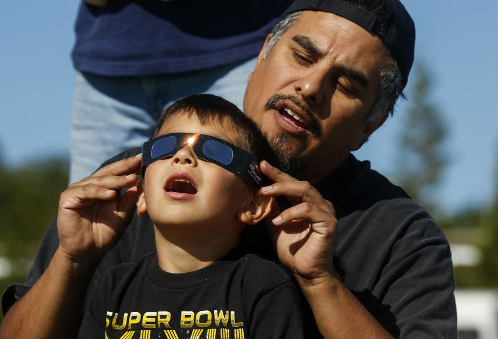 Just off Paine Field near the Future of Flight Aviation Center, Ben Flores of Mukilteo helps his son Vincent, 5, view the solar eclipse through certified safety glasses on Aug. 21. (Dan Bates / The Herald)
