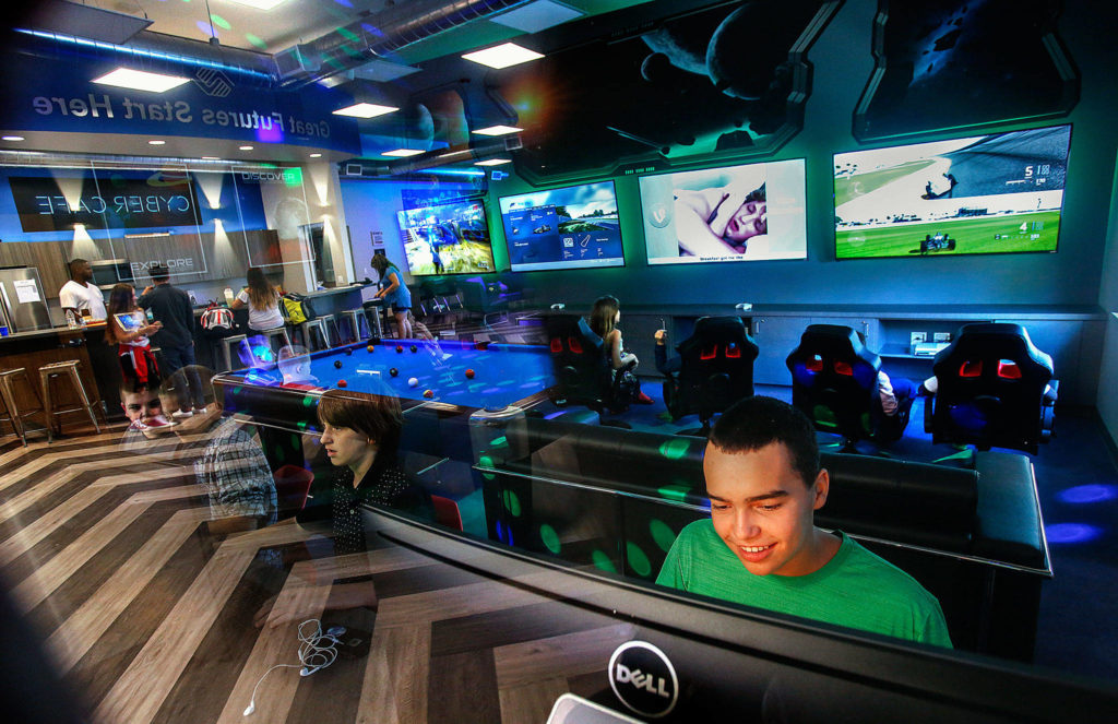 At the new Teen Center at Lake Stevens Boys & Girls Club, Josiah Perez, 12 (green shirt) sits playing a video game in the lower right corner of a menagerie created by reflections and shadows on the plate glass window separating the video/game room and the general purpose room with a pool table, food bar (Cyber Cafe), stools and the people using them. Helping the kids at the cafe is Teen Director Ron Sarrys (left). (Dan Bates / The Herald)
