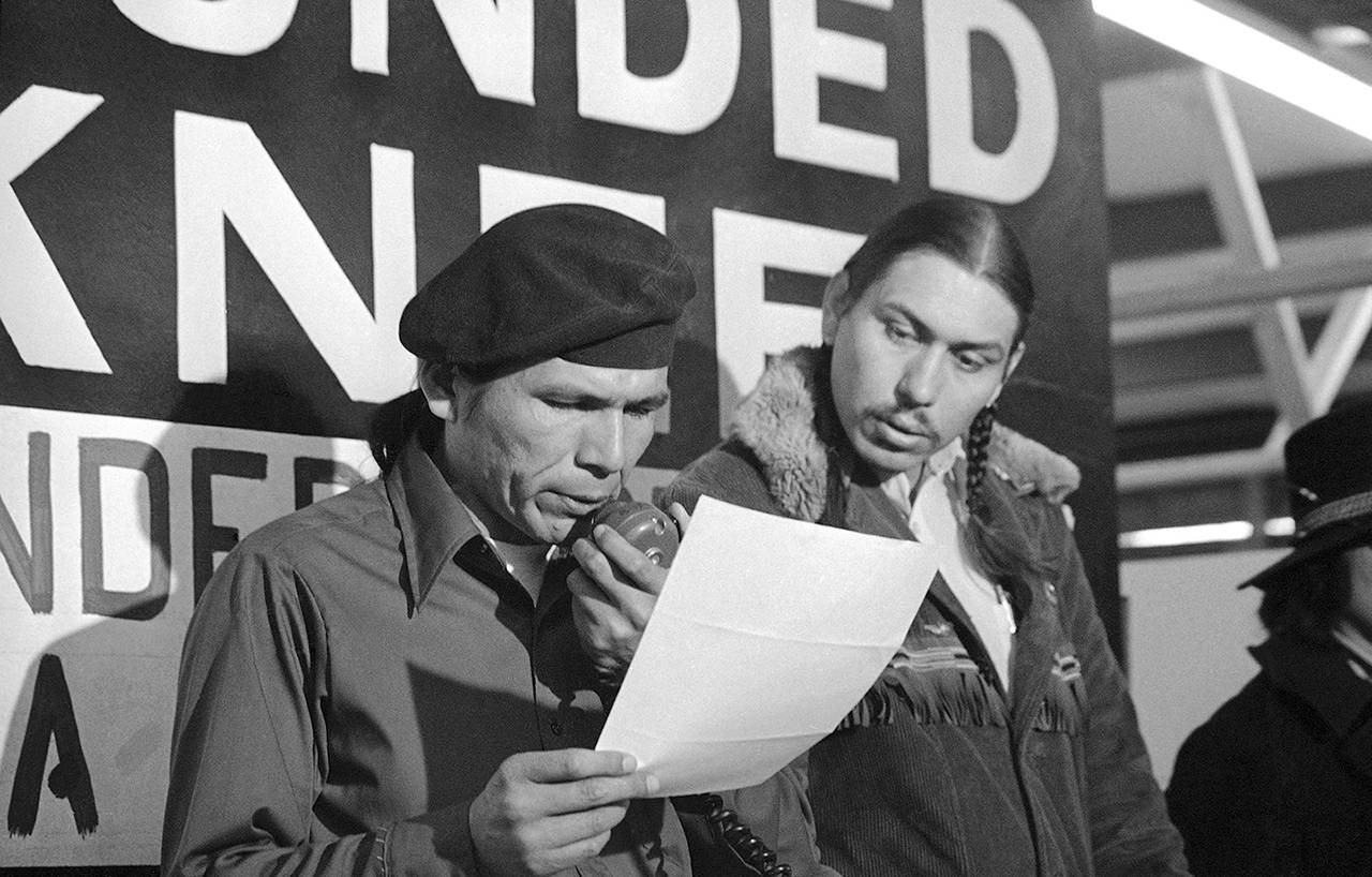 In this 1973 photo taken in Wounded Knee, South Dakota, American Indian Movement leader Dennis Banks (left) reads an offer by U.S. government seeking to effect an end to the Native American takeover of Wounded Knee. Looking on is AIM leader Carter Camp. (AP Photo/Jim Mone, File)