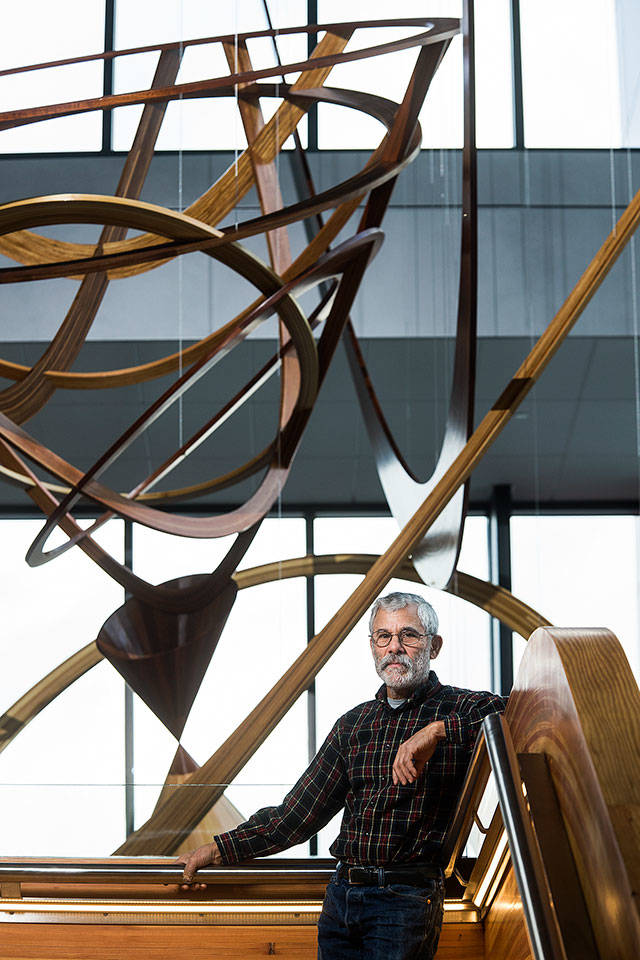Artist Paul Vexler, from Machias, stands the staircase at Washington State University-Everett with his creation behind him. The hanging woodwork art is 35 feet tall and hangs in the main lobby at the school. (Andy Bronson / The Herald)