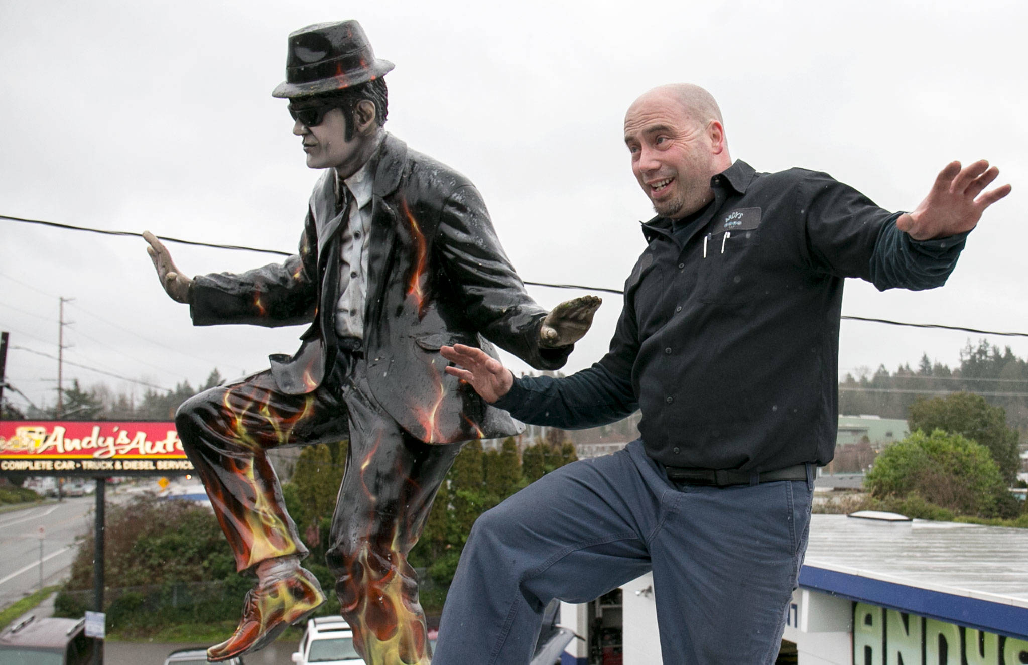 Corey Jonson, owner of Andy’s Auto Repair, strikes a pose with a Blues Brothers figure on the roof of his Lynnwood shop. (Kevin Clark / The Herald)