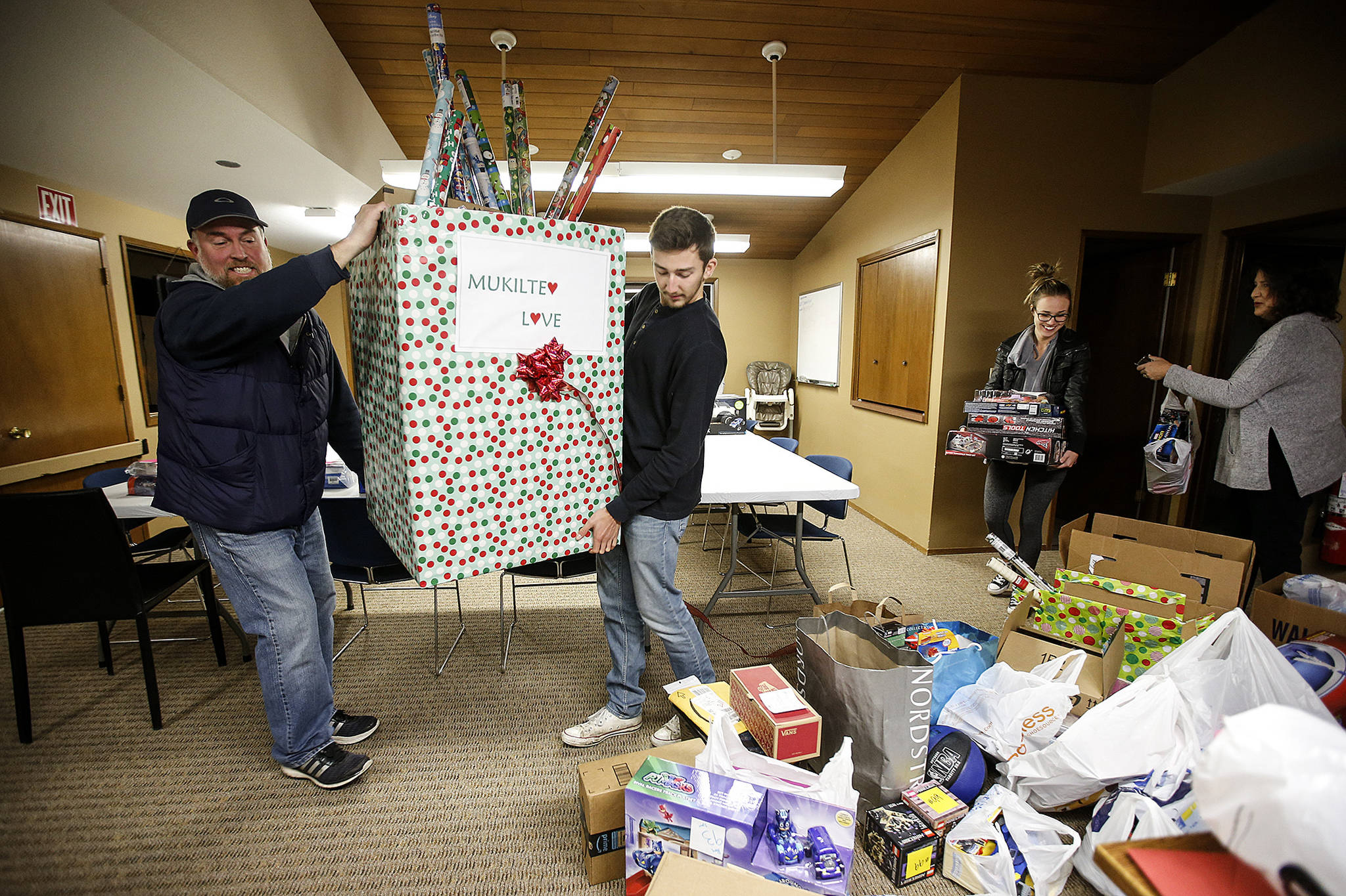 Jason Swift (left) carries a box full of donated presents with Brandon Wright as fellow volunteers Christina Thomas and Jamie Wright help organize other gifts at the Mukilteo Chamber of Commerce building on Tuesday, Dec. 19. The program, Mukilteo Love, was started by Shana Swift and is expected to help provide gifts to over 60 local children in need this holiday season. (Ian Terry / The Herald)