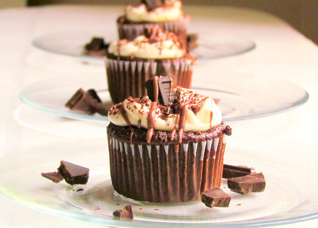 Mini chocolate and cream cakes are topped with cream frosting, dark chocolate chunks and a drizzle of chocolate syrup.

