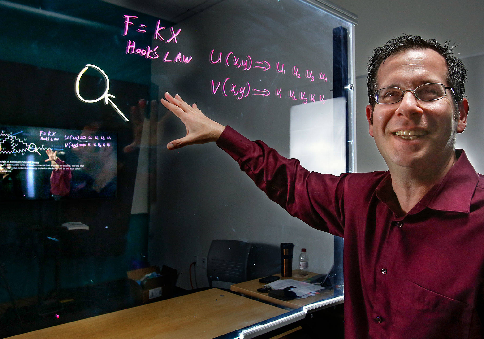 In a lightboard studio, WSU Everett professor Gordon Taub pre-records lectures so he can help students one on one if they need it. He also uses a lightboard in his lectures. It is essentially a large glass surface that he can write and draw on as he speaks face to face with students, as opposed to using a chalkboard with his back to them. It is specifically lit and mounted so content easily stands out against a black background. The video image is flipped horizontally so the symbols and text are not backwards. The system will also allow computer data, photos or pages to share space on the screen with hand-written or drawn content. In the image shown, professor Taub can see the large display screen as it records his teaching efforts.