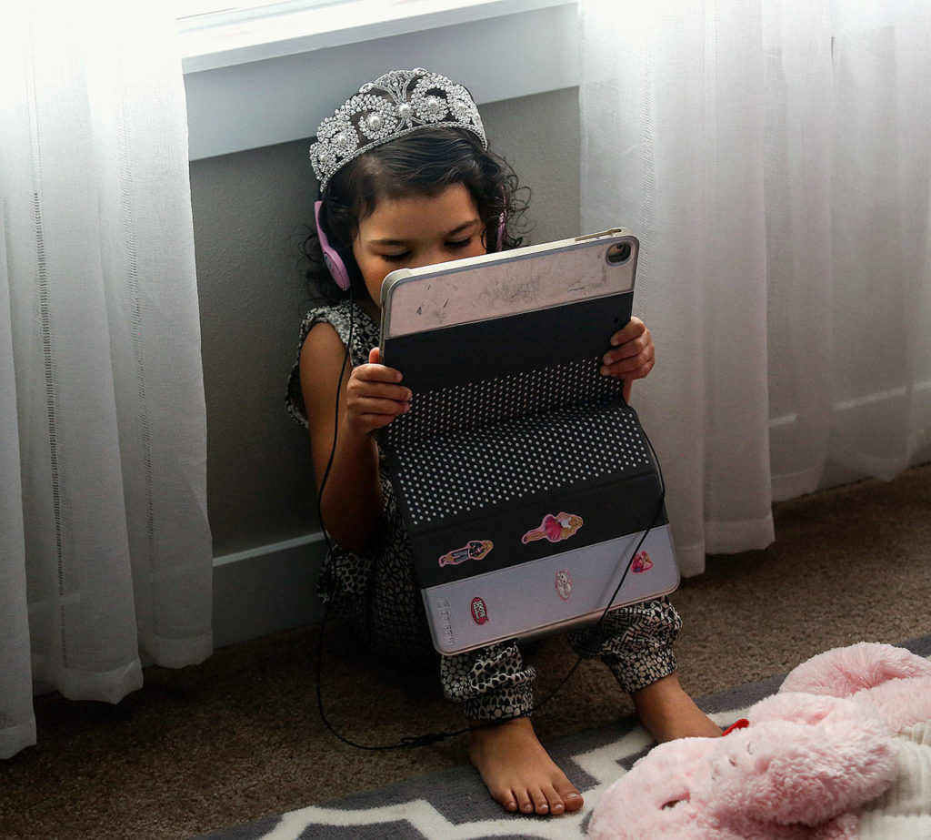 Tiny princess Aria Hansen, 4, appears quite comfortable wearing earphones and her tiara as she sits below a window with an electronic tablet. (Dan Bates / The Herald)

