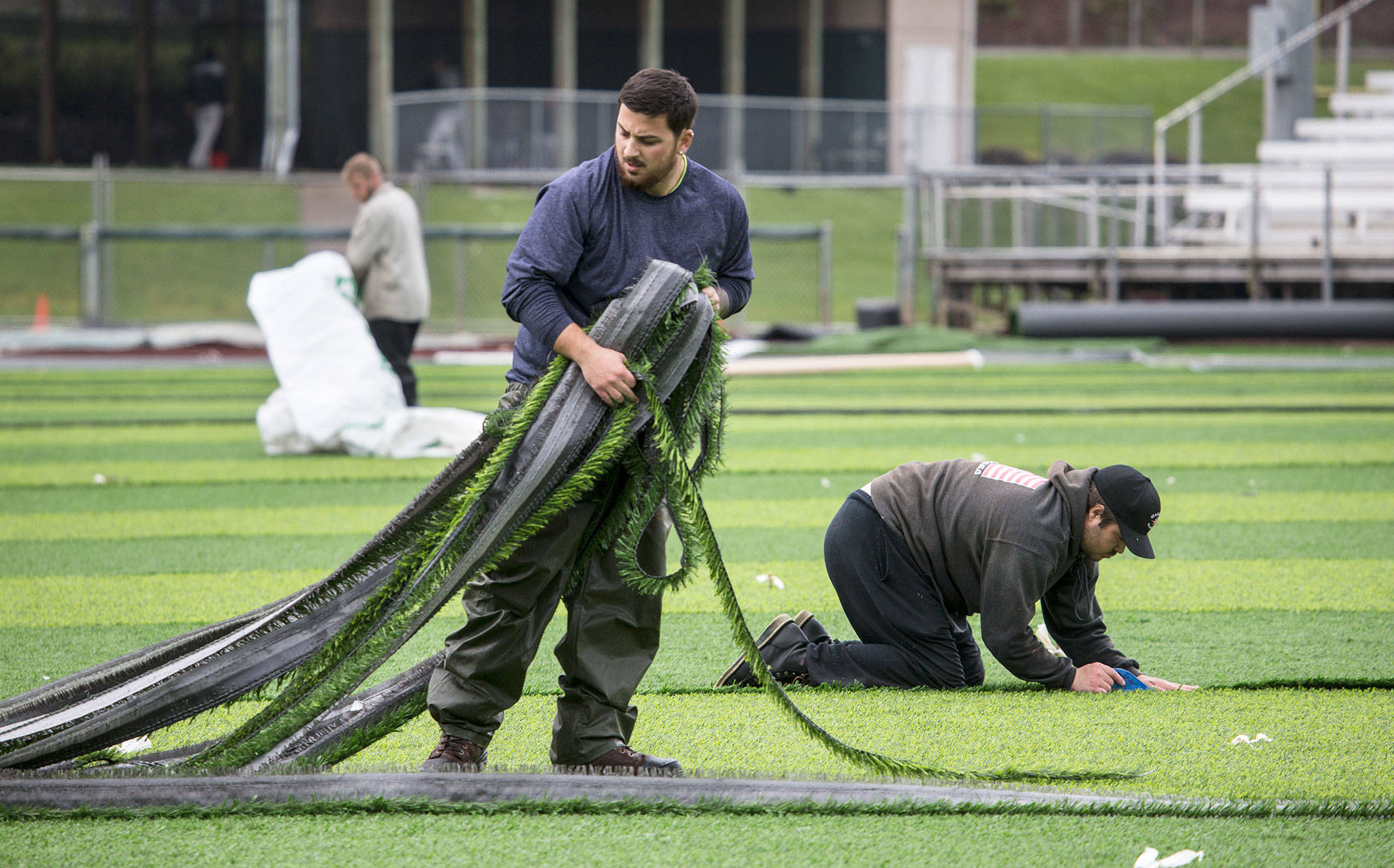 Coast to Coast Turf employee Isaiah Nogueira (center) pulls up turf cut by Tyler Kellogg (right) as Justin Conner Buchanan bags scraps. The three men were part of crew installing a new turf field at Everett Memorial Stadium on Friday. (Andy Bronson / The Herald)