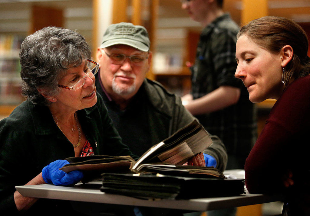 The Everett Public Library’s “History-Mystery” night drew a number of photo sleuths eager to look at old scrapbooks and try to identify people and places from the past. Here, Judy Brackett (left) and Steve Hudspeth look over some very old scrapbook items as the library’s Northwest Room historian, Lisa Labovitch (right), looks on. (Dan Bates / The Herald)
