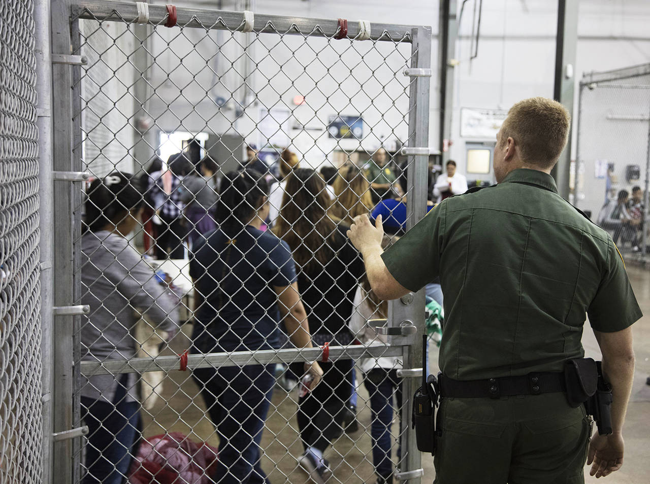 A U.S. Border Patrol agent watches as people who’ve been taken into custody related to cases of illegal entry into the United States stand in line at a facility in McAllen, Texas, on Sunday. (U.S. Customs and Border Protection’s Rio Grande Valley Sector via AP)