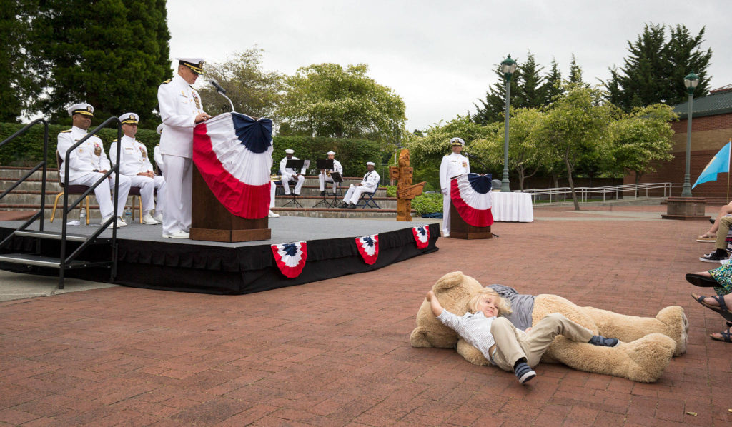 Capt. Mike Davis, the new commanding officer of Naval Station Everett, speaks to the crowd as his son, Liam Davis, 3, plays with giant teddy bear during the Change of Command Ceremony on Friday in Everett. (Andy Bronson / The Herald) 
