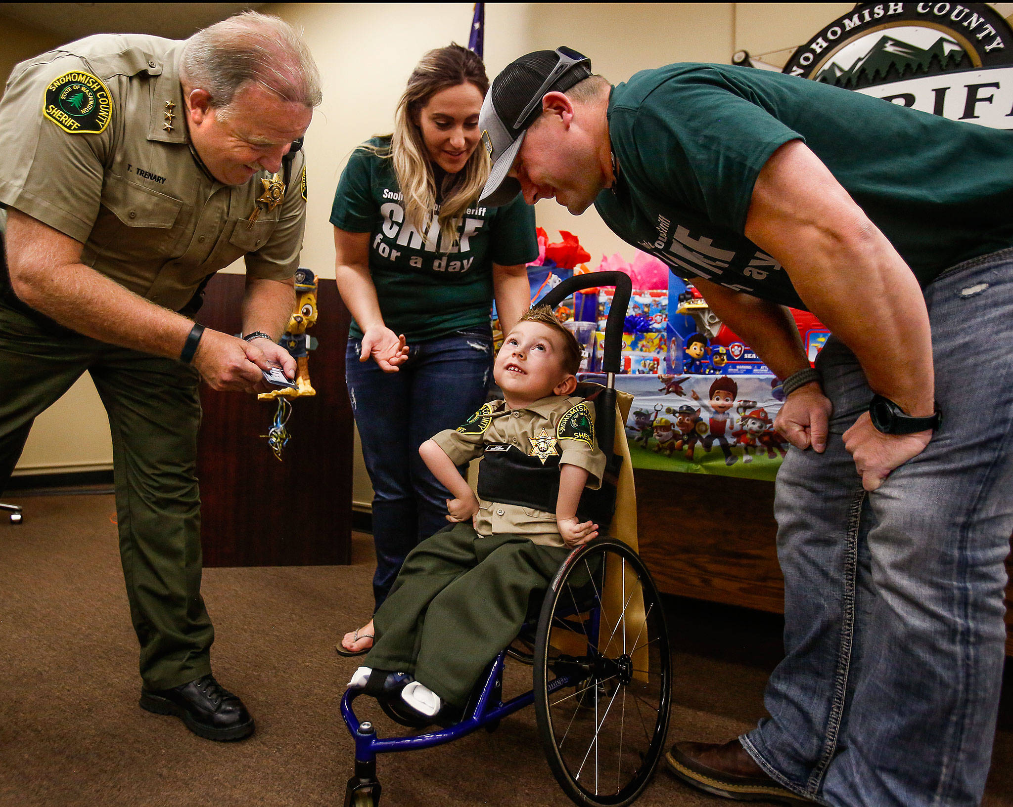 From his wheelchair, Isaiah Kobernik, 4, looks up happily as his parents, Gerad and Anastassia Kobernik, and Snohomish County Sheriff Ty Trenary lean over him after his dad pinned a shiny new sheriff’s badge on his shirt following a presentation to him by Trenary at the Snohomish County Courthouse on Tuesday. Isaiah has spinal muscular atrophy, which affects the motor nerve cells in the spinal chord. (Dan Bates / The Herald)