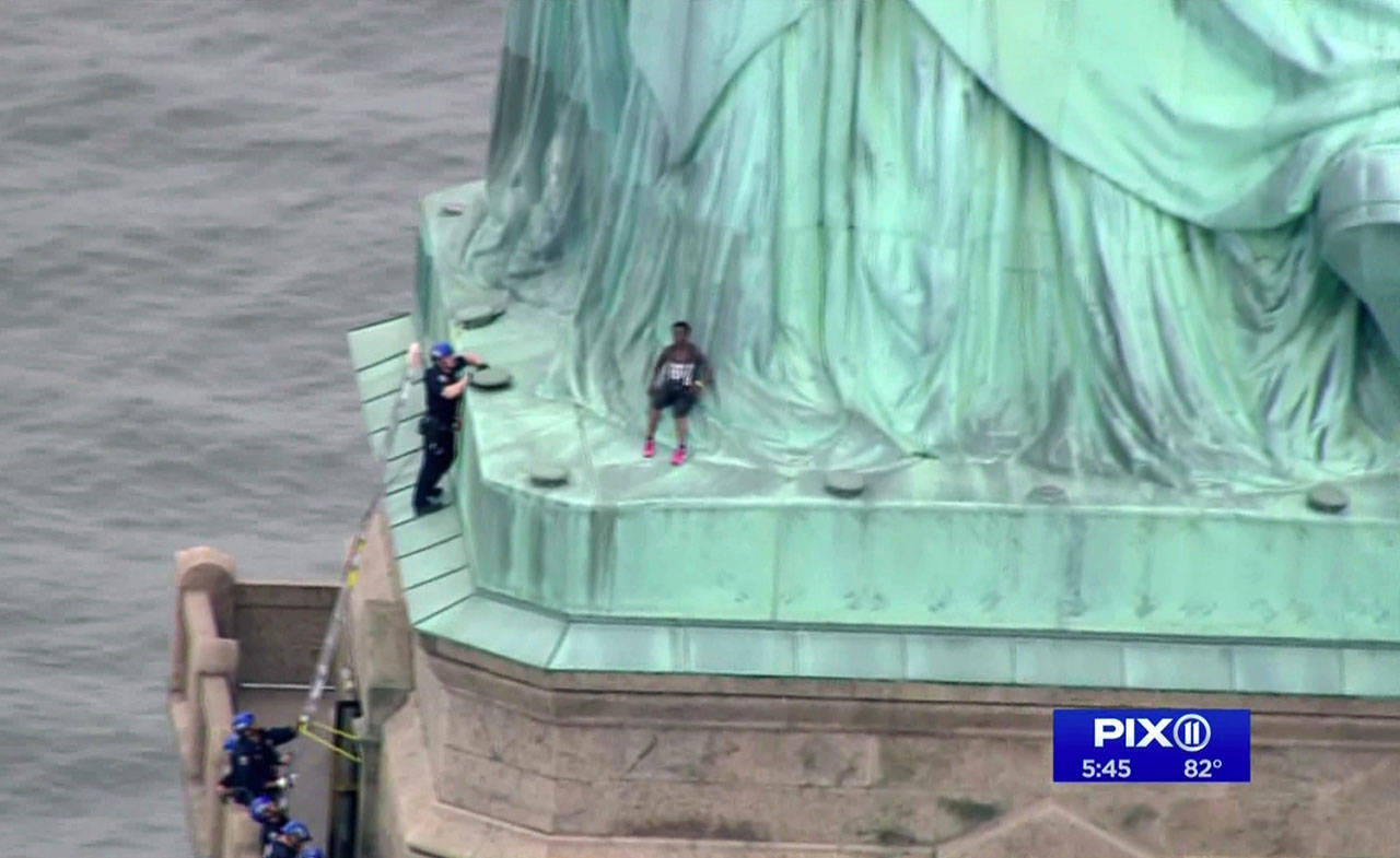 In this image a person (center) leans against the robes of the Statue of Liberty on Liberty Island, as one of the police officers climbed up on a ladder to stand on a ledge nearby talking the climber into descending in New York on Wednesday. (PIX11 via AP)