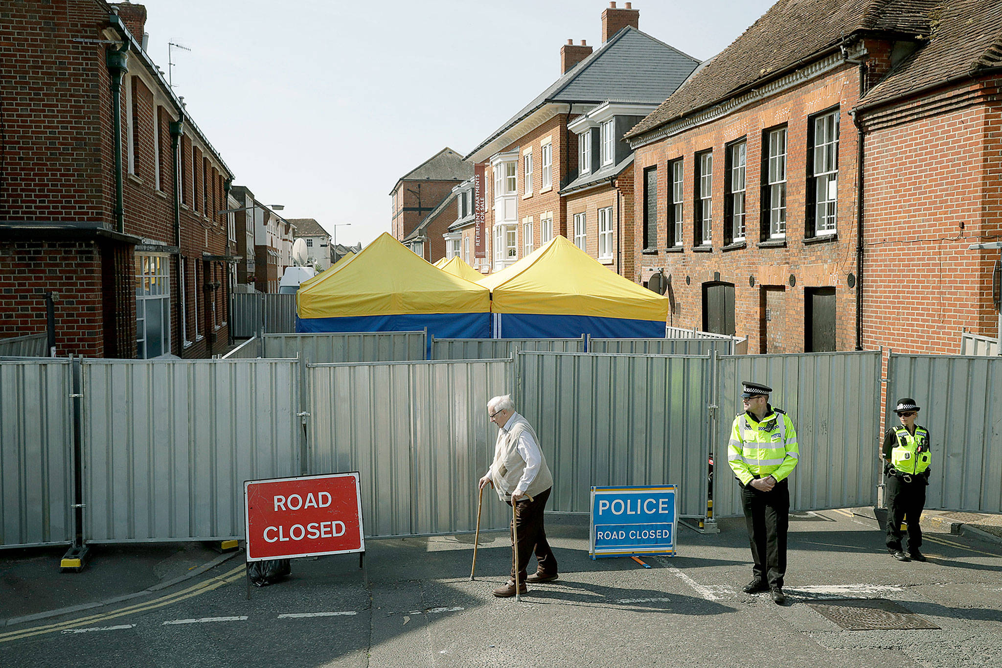 A man walks by as British police officers guard metal fencing surrounding tents set up by search teams at the end of Rollestone Street, outside the location of the John Baker House for homeless people in Salisbury, England. British detectives investigating the poisoning of two people by the nerve agent Novichok in southern England say scientists have found the source of the deadly substance. (AP Photo/Matt Dunham, File)