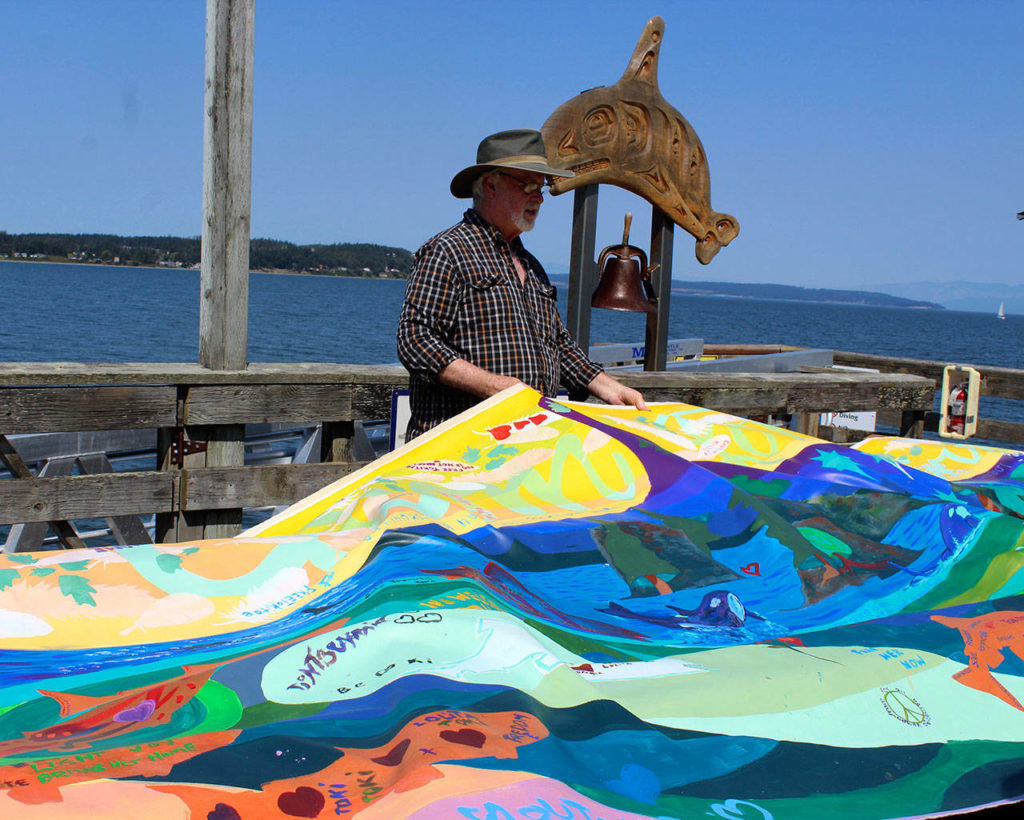 On the Coupeville wharf near the whale bell, Rick Baker helps unfurl a banner painted along the route of the Tokitae Totem Pole Journey undertaken by the Lummi Nation in May. (Patricia Guthrie/Whidbey News-Times)
