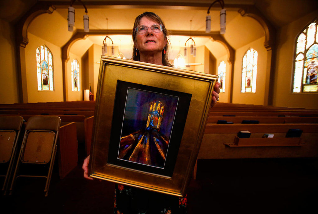 Holding one of several paintings she created and brought in for the Everett Art Walk, Laurie Povey Crawford looks up at the stained-glass window facing east in the First Presbyterian Church on Thursday night. Crawford lived in Everett 45 years before learning from a Herald article that those spectacular windows had been created in 1910 by the Povey Brothers Studio, her great-grandfather’s business in Portland. The painting she holds portrays streams of light from that stained glass window. (Dan Bates / The Herald)
