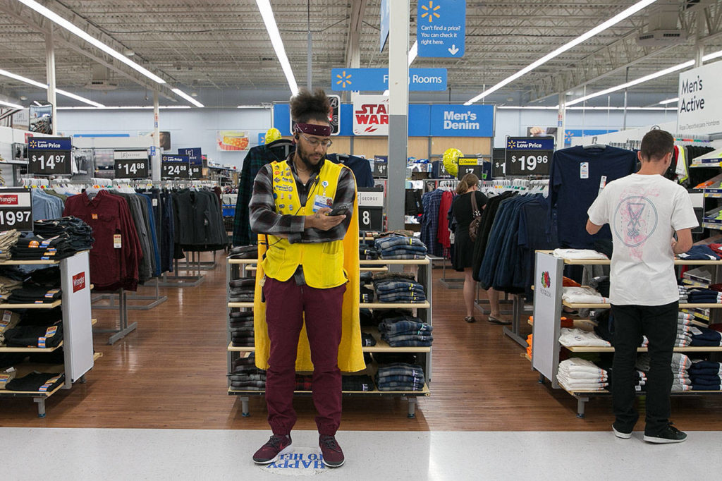 Isaiah Owens awaits a customer service call from his “happy to help” spot on the floor of the Everett Walmart. He began wearing a cape as part of a 2017 back-to-school seasonal promotion and never took it off. (Kevin Clark / The Herald) 
