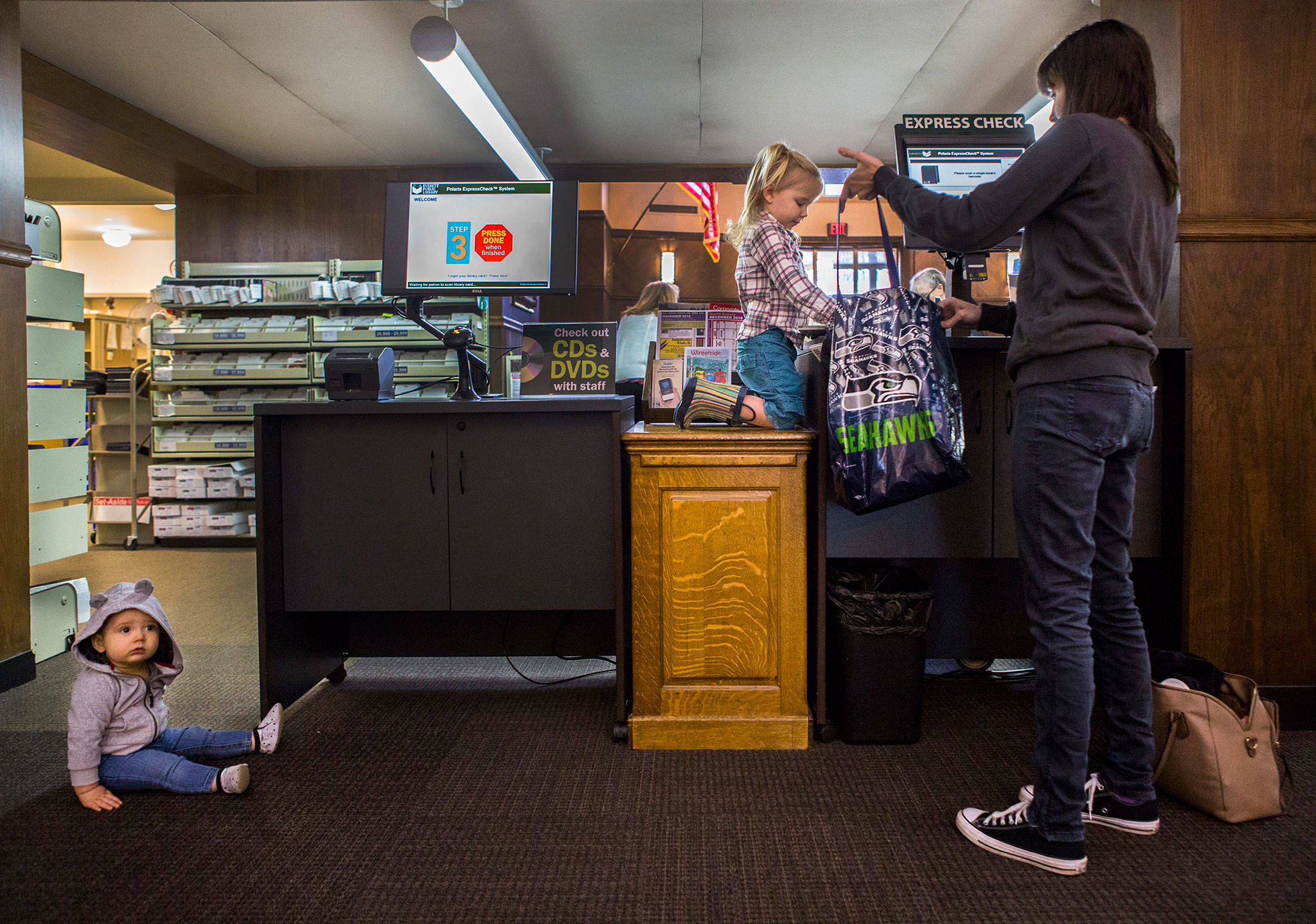 Rachel Hawkins (right) holds open a book bag for her daughter, Talia Hawkins, 3, while brother Weston Hawkins, 9 months, waits in the Everett Public Library on Wednesday. The city is looking to reduce costs in library, fire and transit services next year. (Olivia Vanni / The Herald)