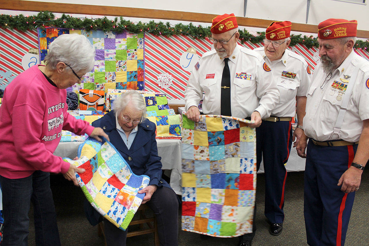 Pam Donery admires quilts made by Ginny Mayer as Marine Corps veterans Frank Thornton, Ed Donery and Tom Keltner thank her for the donation to the Toys for Tots program. (Patricia Guthrie / Whidbey News Group)