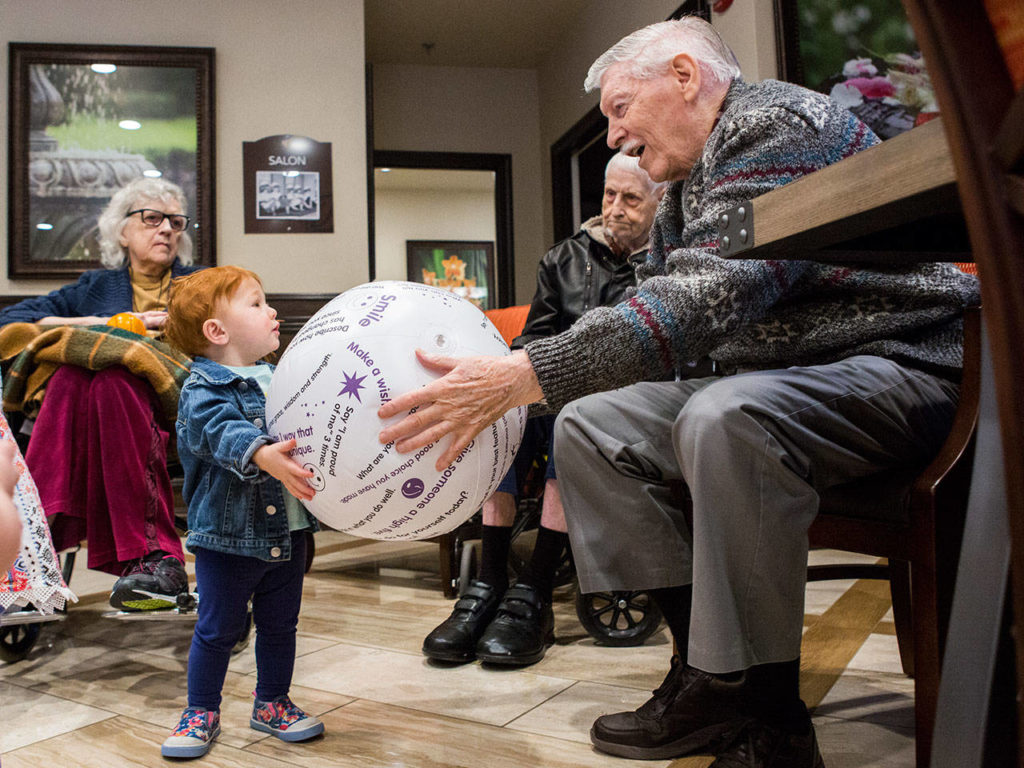 Ralph Oberstadt, 88, hands a beach ball to Alexia Klauber, 1, during the Seniors & Sprouts program at Pacifica Senior Living in Lynnwood. (Olivia Vanni / The Herald)
