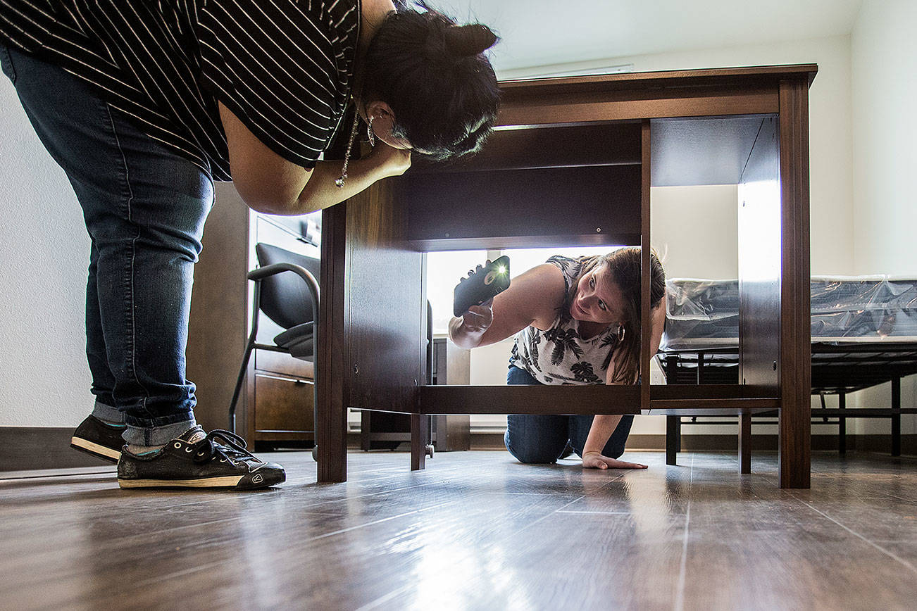 Premera Blue Cross volunteer Jaimie Powell uses her cell phone light to look for a hole in a desk, as she and Michelle Rosenblad assemble furniture at the new Cocoon House on Colby Avenue on April 10. (Andy Bronson / The Herald)