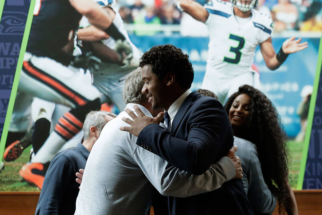 Seattle Seahawks quarterback Russell Wilson, center, hugs head coach Pete Carroll, left, in front of a photograph of Wilson in action, as Wilson’s wife Ciara looks on at right, Wednesday, April 17, 2019, prior to a NFL football press conference in Renton, Wash. Earlier in the week, Wilson signed a $140 million, four-year extension with the team. (AP Photo/Ted S. Warren)