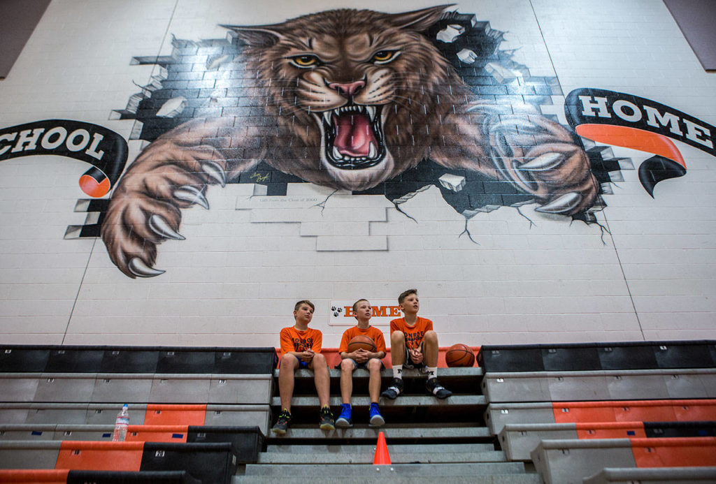Three 12-year-olds — AJ Welch (left), Myles Baumchen (center) and Wyatt Prohn — sit under the Bearcats logo and watch a scrimmage during a youth basketball camp Wednesday at Monroe High School. (Olivia Vanni / The Herald)
