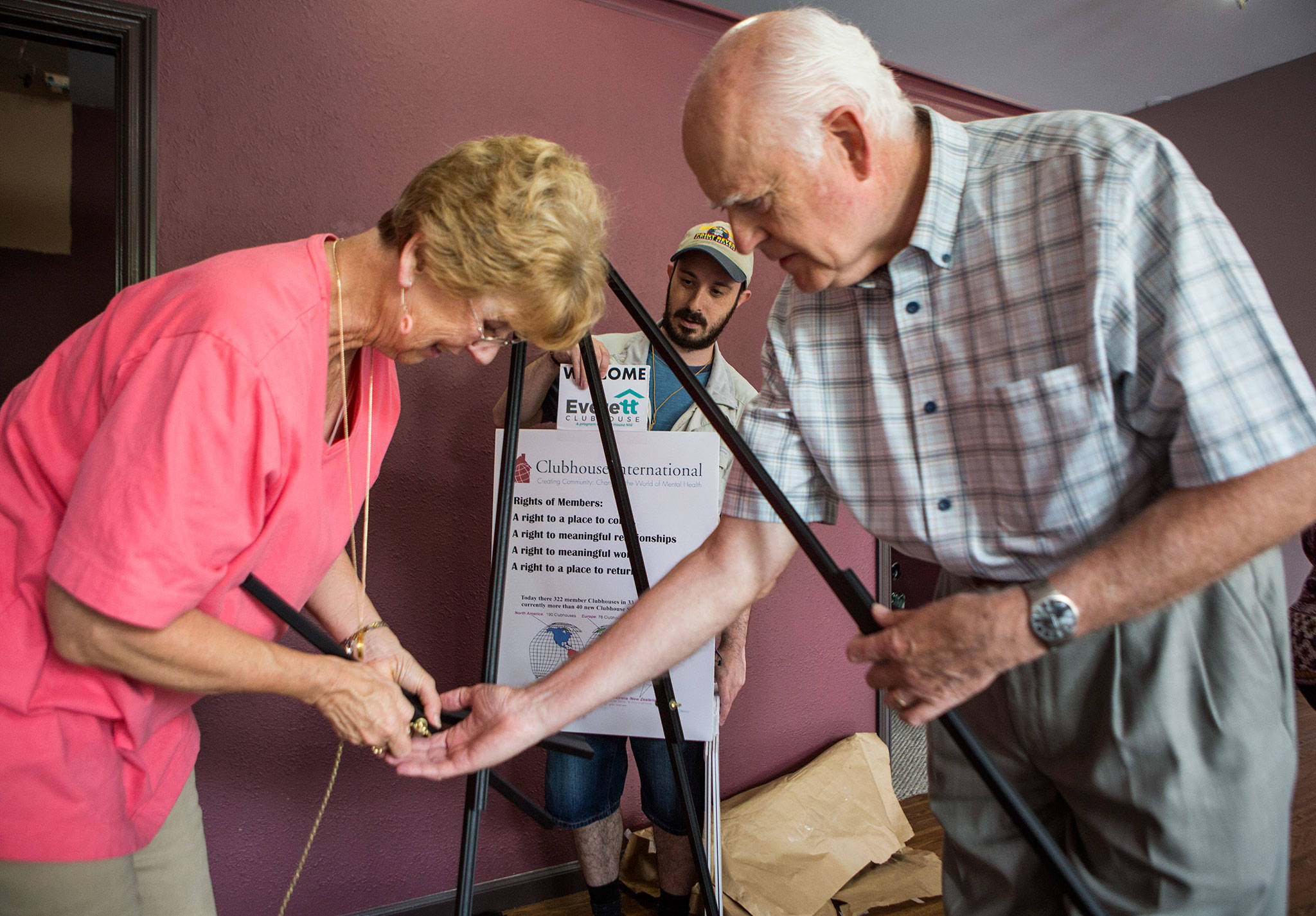Clubhouse member Alex Odesskiy (center) helps Meg McClure (left) and Meg’s husband, Harold McClure (right), set up a poster stand at the Everett Clubhouse. Harold McClure calls the opening of the clubhouse “the fulfillment of a vision.” (Olivia Vanni / The Herald)