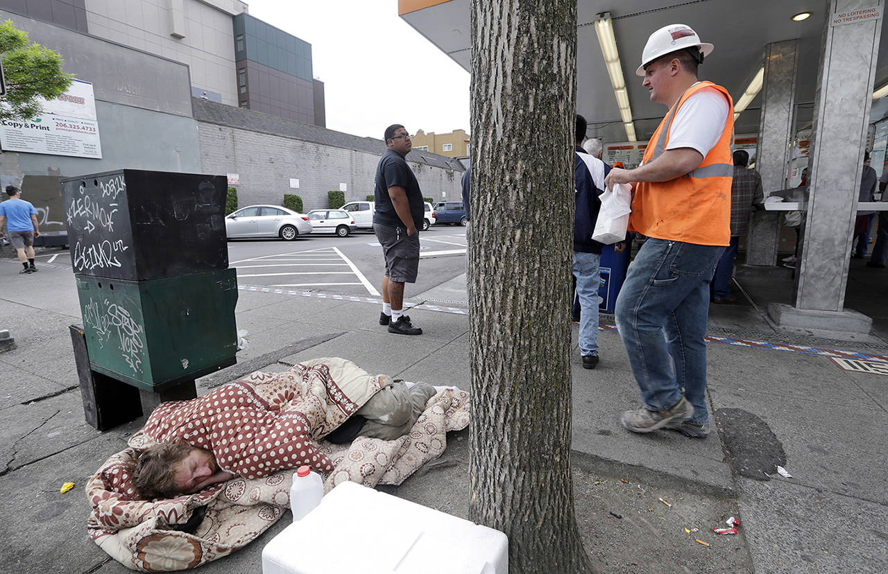 In this May 2018 photo, a man sleeps on the sidewalk as people behind line up to buy lunch at a Dick’s Drive-In restaurant in Seattle. (AP Photo/Elaine Thompson, File)