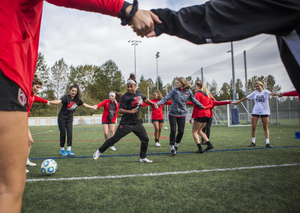 Everett Community College’s Olivia Lee (left) and Elizabeth Erickson hold hands in the center of a circle during a passing drill during the Trojans team practice on Oct. 8 at Kasch Park in Everett. (Olivia Vanni / The Herald)
