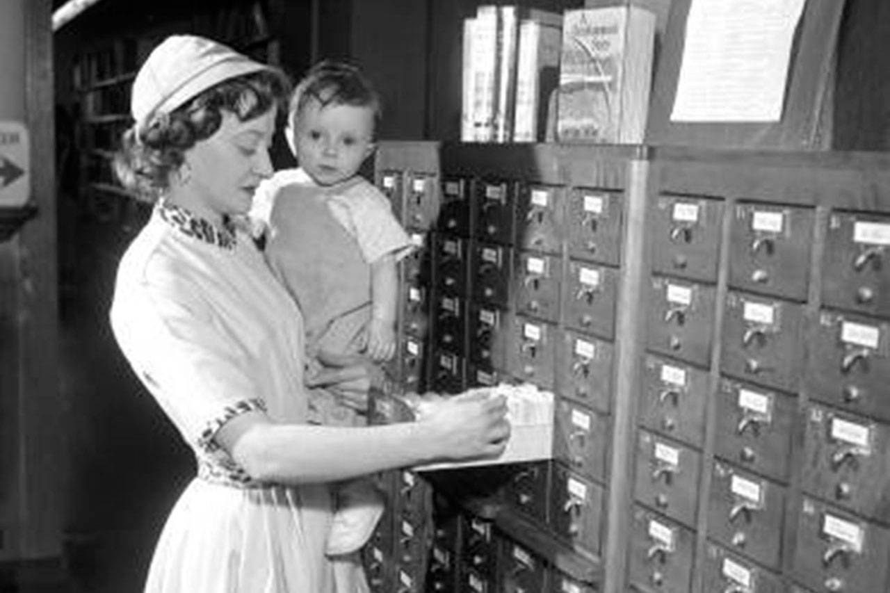 A woman, holding a child, looks through book titles in the card catalog at Everett Public Library in 1956. E-books now allow library users to download books, but one publisher recently changed its rules for libraries, curtailing the availability of e-books. (Everett Public Library Northwest Room Collection)