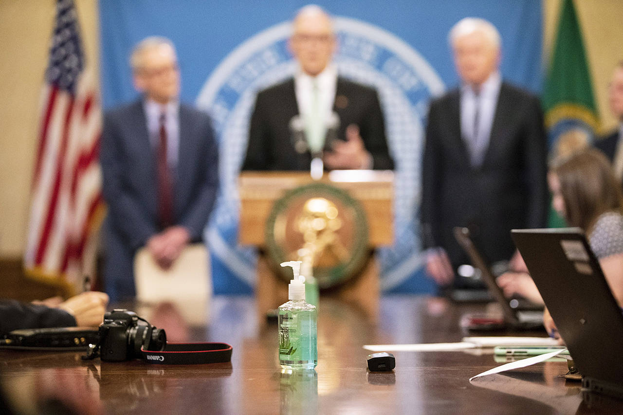 A bottle of hand sanitizer sits in front of reporters as Gov. Jay Inslee gives updates on COVID-19 cases in Washington state on Monday in Olympia. The governor was joined by Dr. John Wiesman (left), secretary of the Washington State Department of Health and Robert Ezelle (right), director of the Washington State Department of Emergency Management. (Amanda Snyder/The Seattle Times via AP)