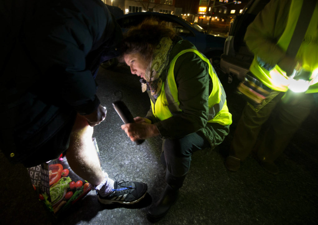 Sheila Murphy, a nurse anesthetist with MercyWatch, looks over a homeless man’s swollen knee in a parking lot near the Everett Public Library on Feb. 18. (Andy Bronson / The Herald)
