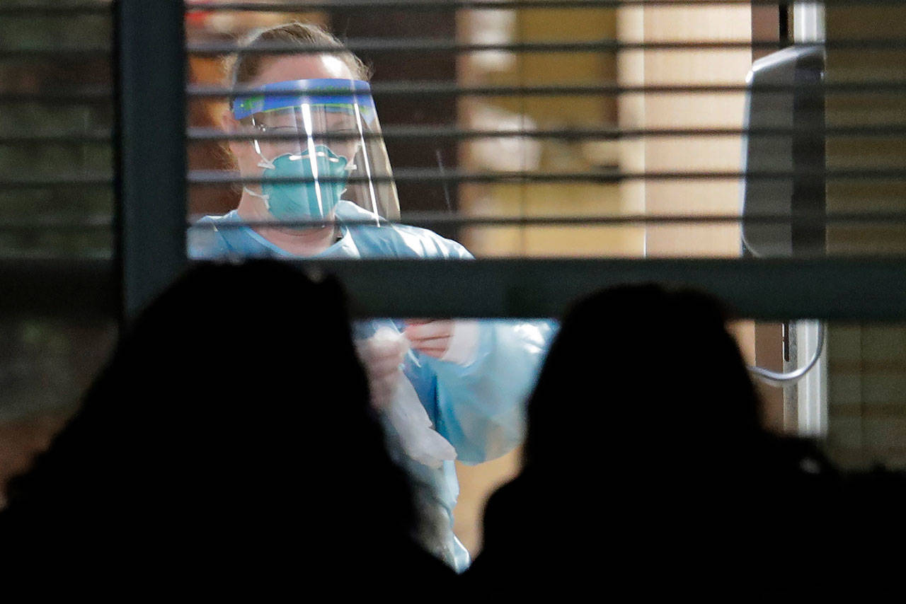 In this March 10 photo, a worker wearing protective gear is seen through a window as she works in a room of a woman who has tested positive for the new coronavirus, as her daughters look in from outside the window, at Life Care Center in Kirkland in King County. Four similar facilities in Snohomish County have confirmed or probable COVID-19 cases. (AP Photo/Ted S. Warren, File)