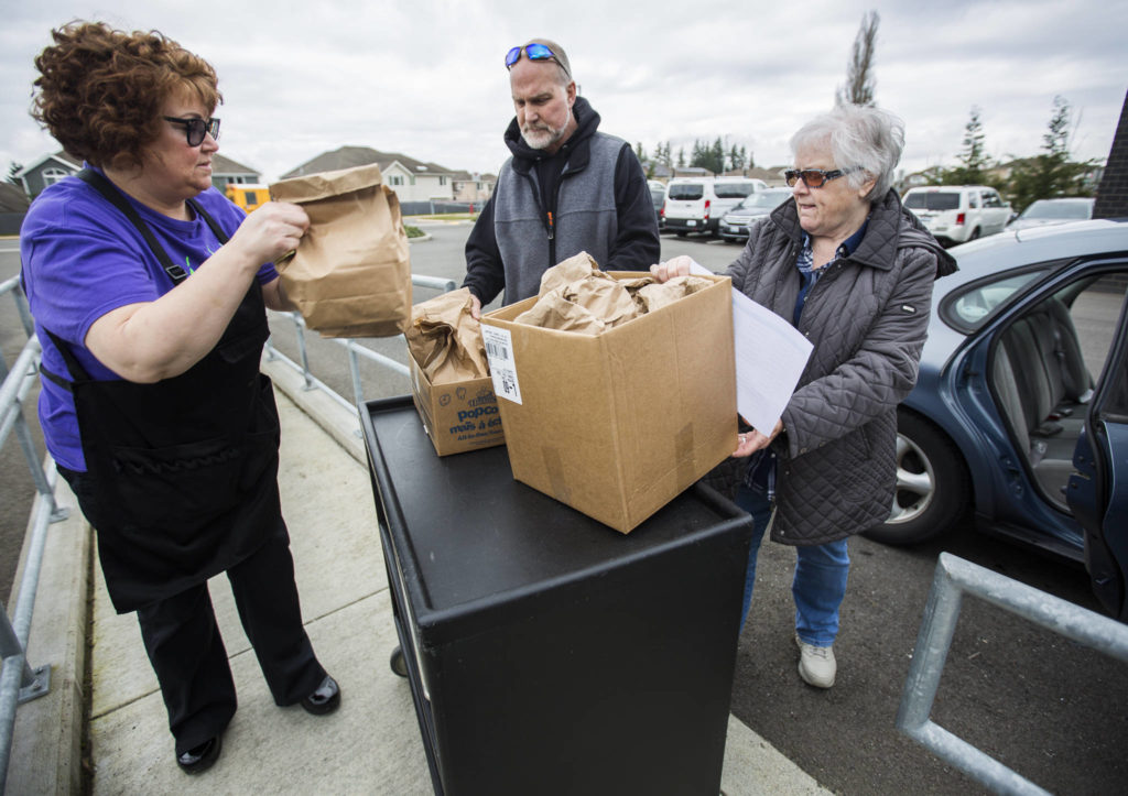 Kitchen manager Kristine Bengochea (left) hands meals to Bruce Kelly (center) and Gail Fox to get delivered on March 12 in Bothell. (Olivia Vanni / The Herald)