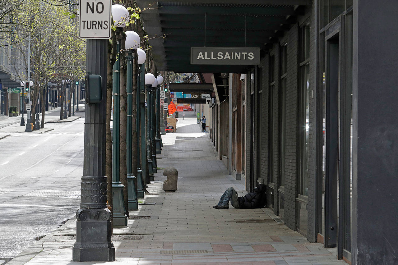 A person sleeps on the sidewalk near a closed AllSaints clothing store Saturday in downtown Seattle. Streets remained mostly empty due to Washington state’s ongoing stay-at-home order and non-essential businesses continuing to be closed as a result of the outbreak of the coronavirus. (AP Photo/Ted S. Warren)