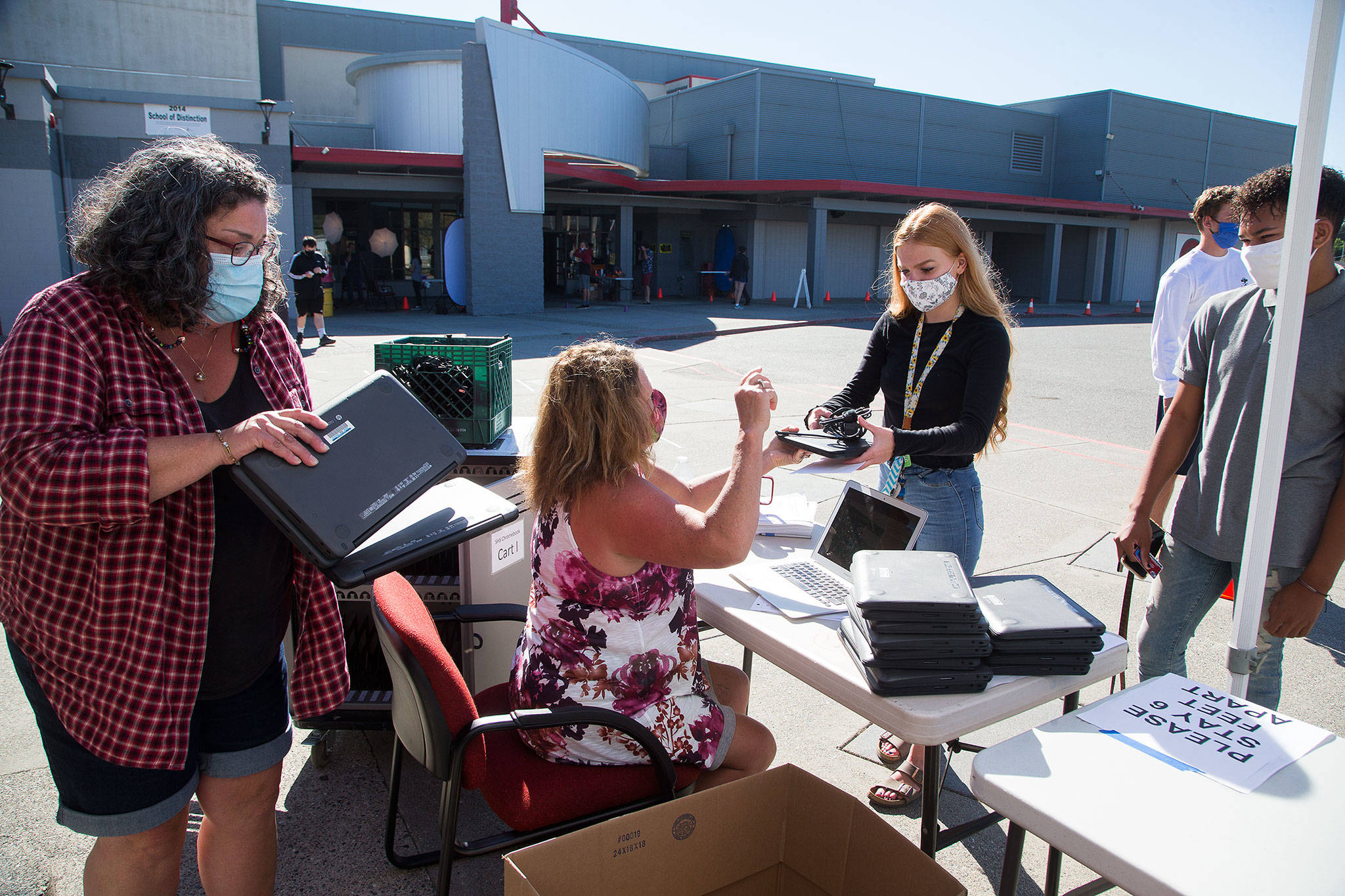 Sheryl Wark stacks computers as Stanwood High’s Renee Herigstad (center) hands a Chromebook to junior Savannah Wickman on Wednesday in Stanwood. Students in the district will be learning online. (Andy Bronson / The Herald)